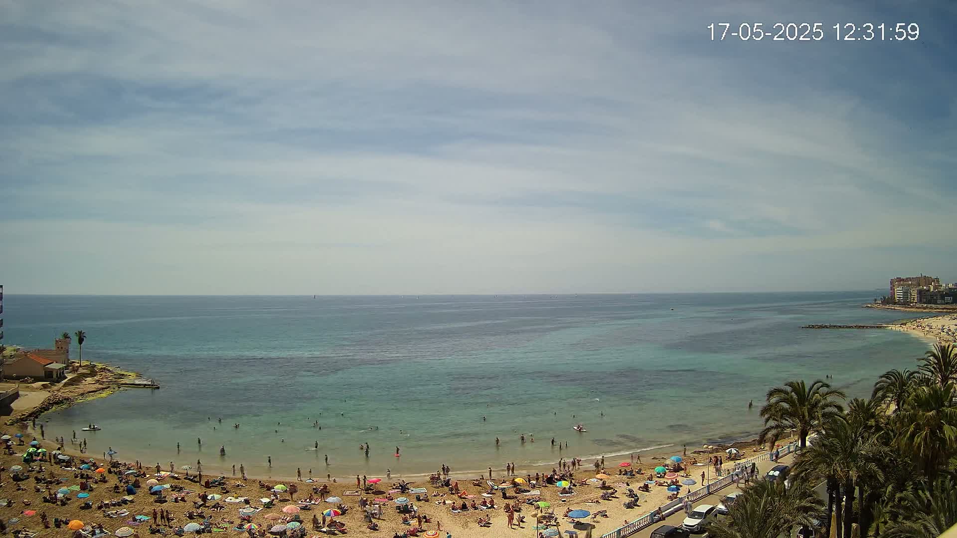 A sandy beach with many sunbathers under colorful umbrellas borders a calm, teal-green sea under a partly cloudy sky.