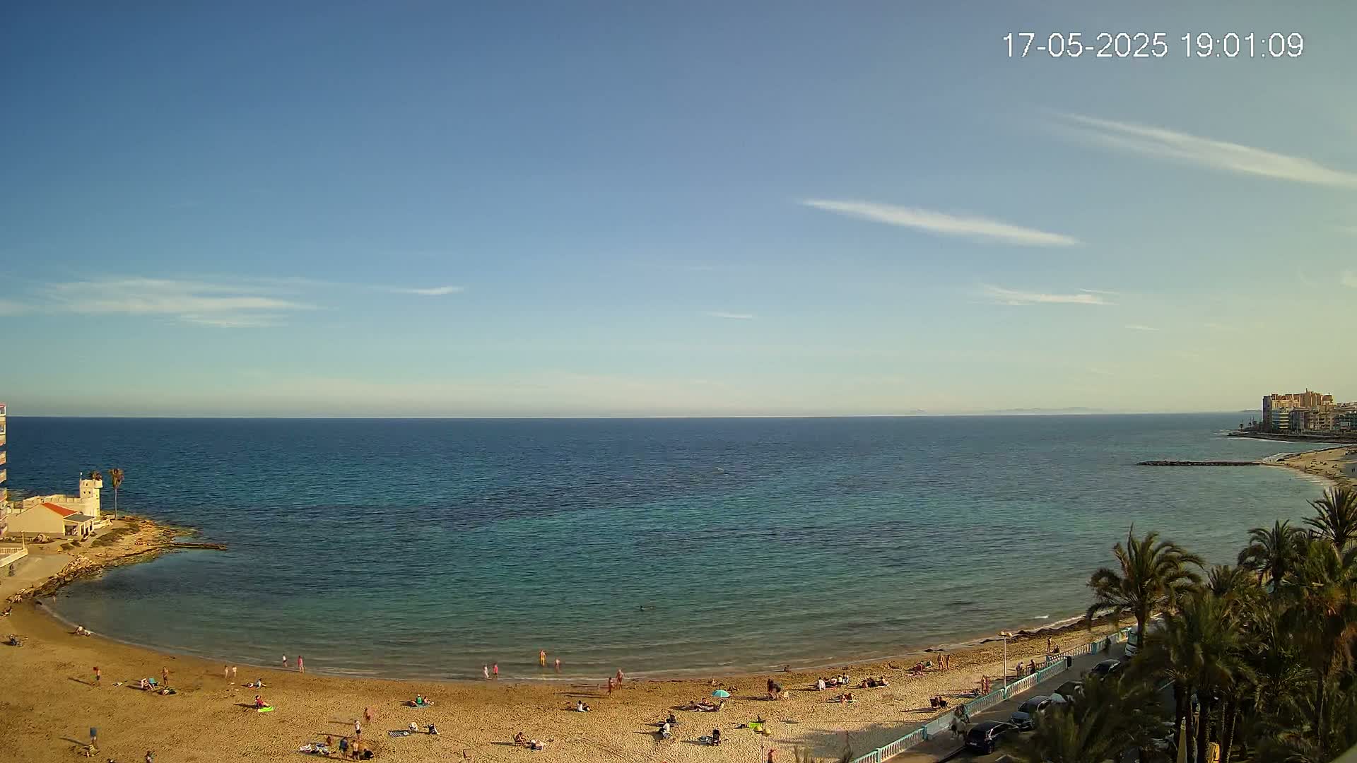 A sandy beach with scattered people is nestled in a cove next to calm, clear blue water under a mostly clear sky.