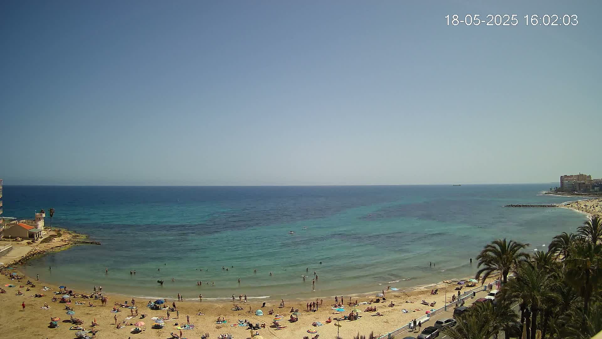 A sandy beach with numerous people sunbathing and swimming in a calm, clear turquoise sea under a bright, sunny sky.