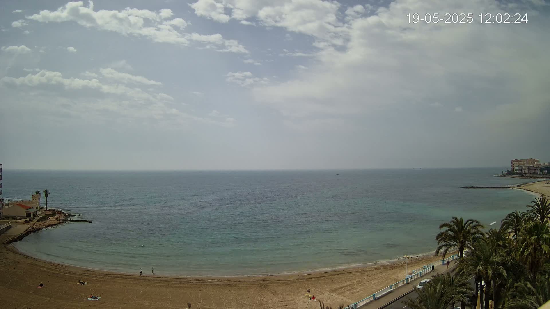 A sandy beach curves along a calm, light-blue sea under a partly cloudy sky.