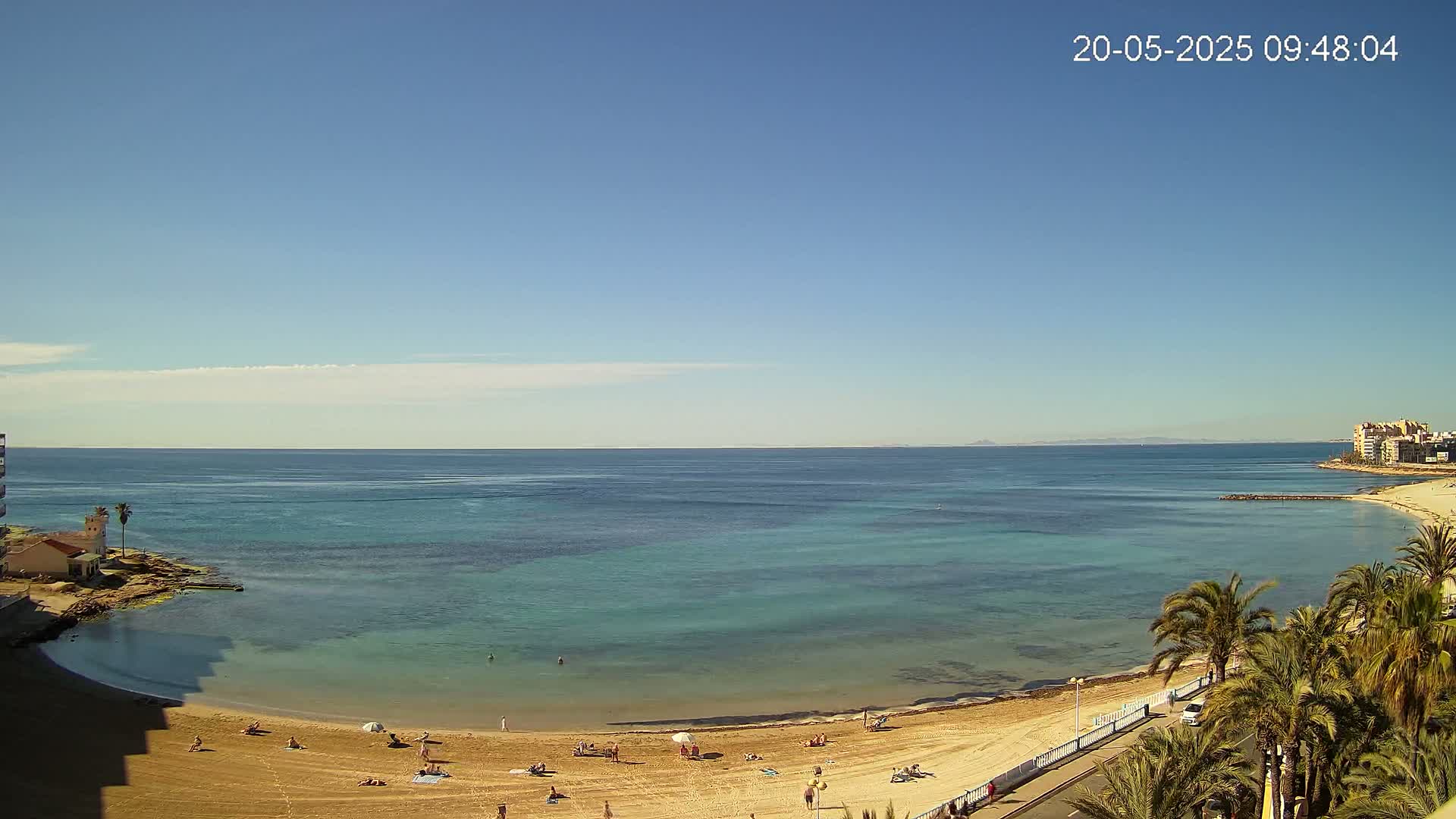 A mostly sunny day at a sandy beach with calm, clear turquoise water, palm trees, and a few people sunbathing.