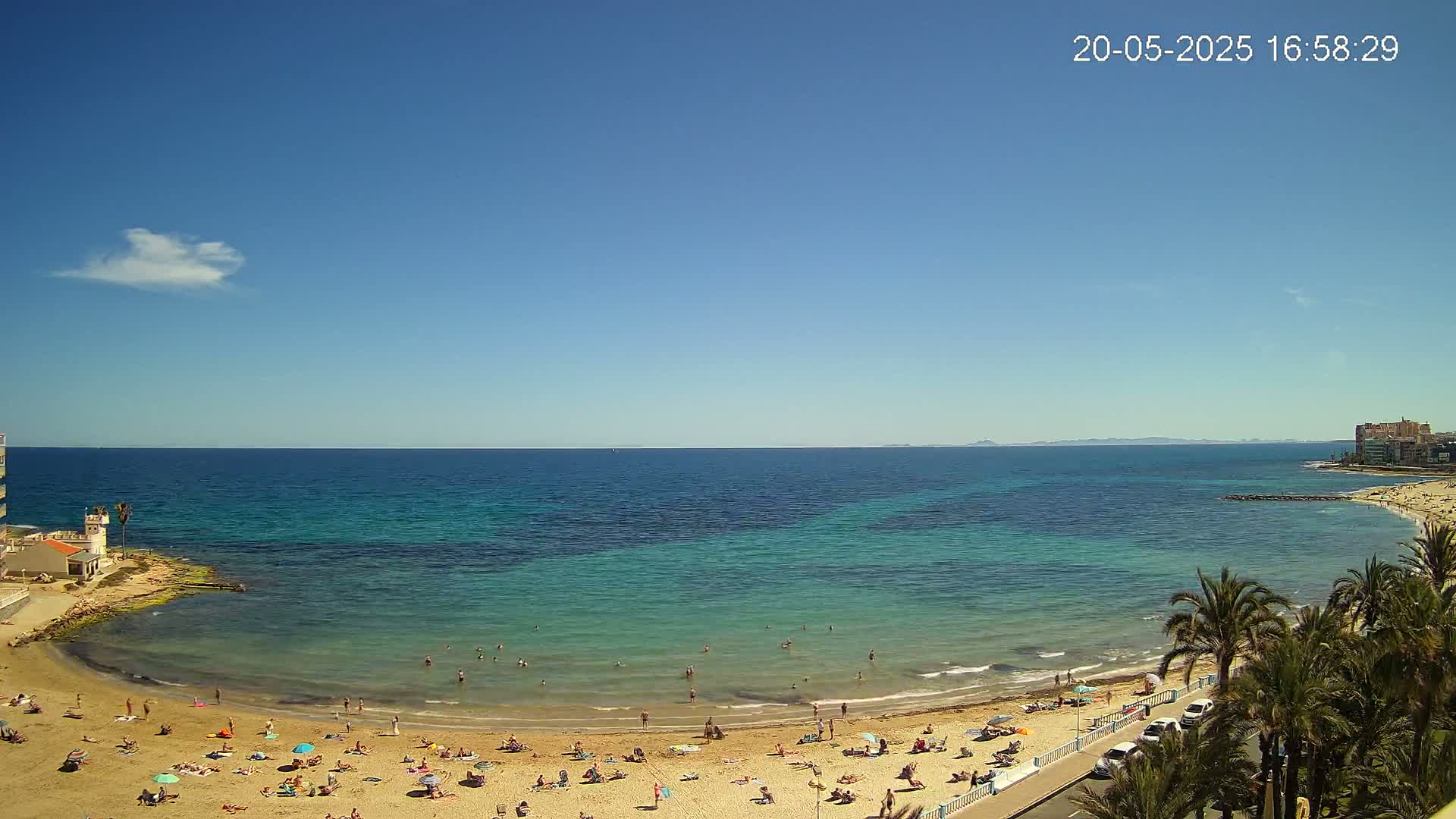 A sandy beach with numerous sunbathers and swimmers is nestled in a cove of clear, turquoise water under a bright, sunny sky.