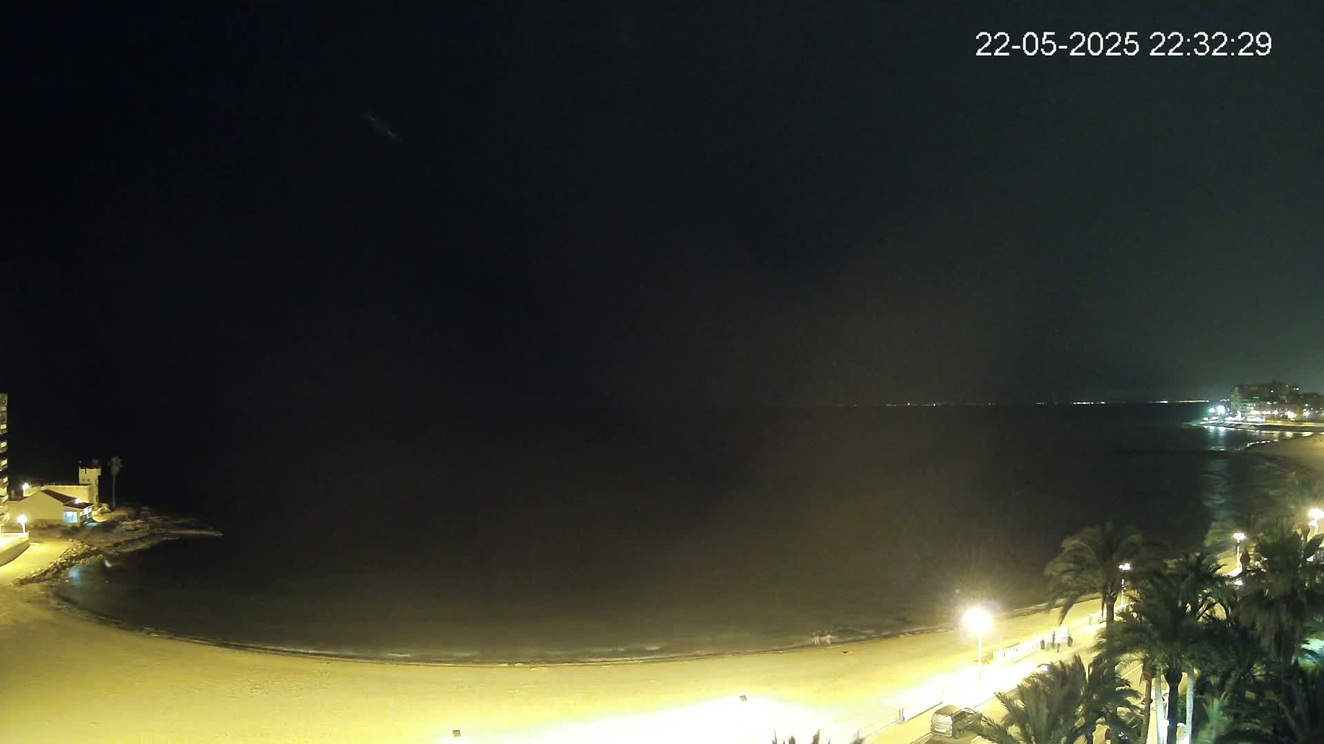 A nighttime view of a sandy beach curving into a calm, dark sea under a clear, dark sky, illuminated by lights along a palm tree-lined walkway.