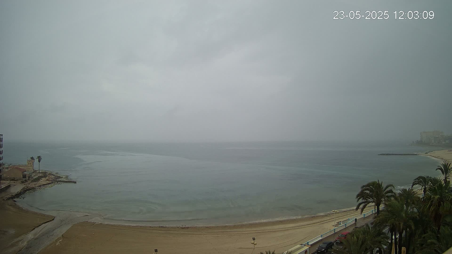 A hazy, overcast day reveals a sandy beach curving into a calm sea, with palm trees and buildings visible in the foreground.