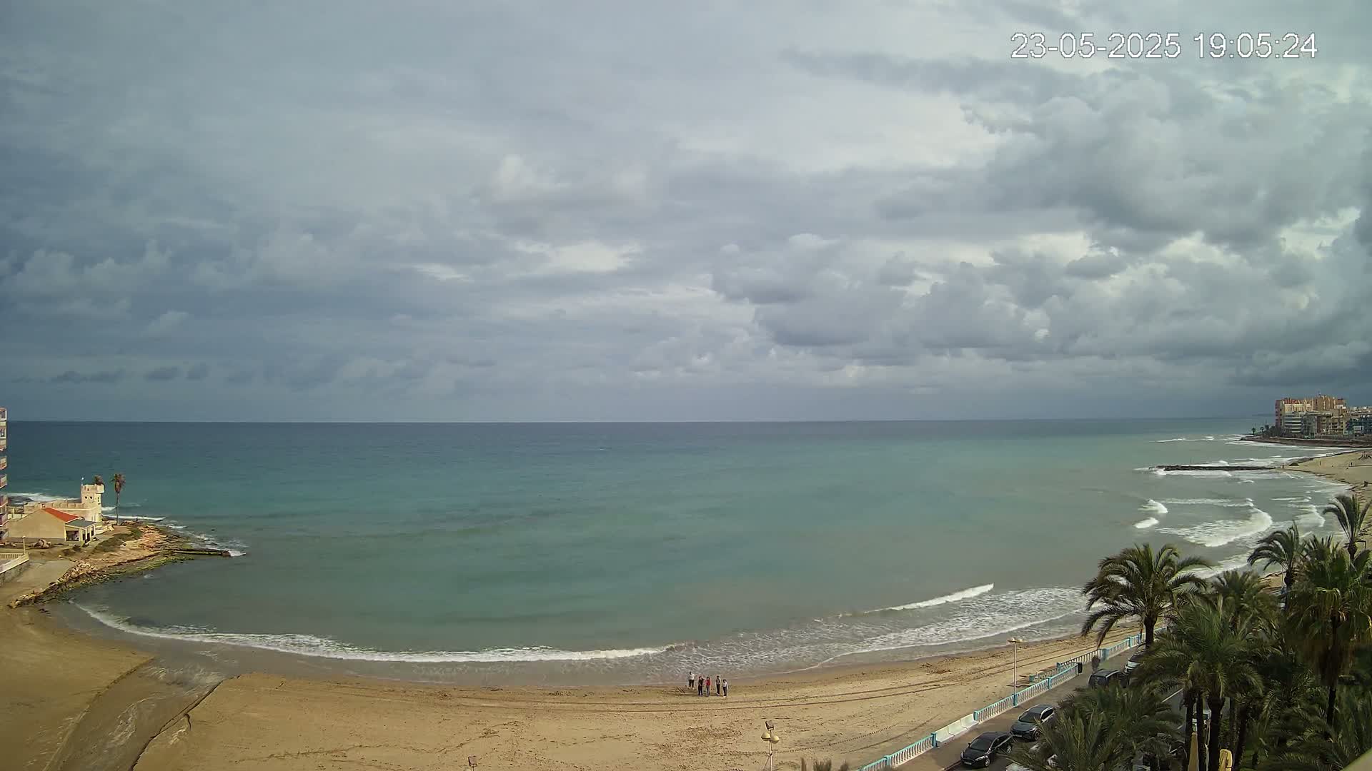 A sandy beach with calm waves laps a turquoise sea under a mostly cloudy sky.