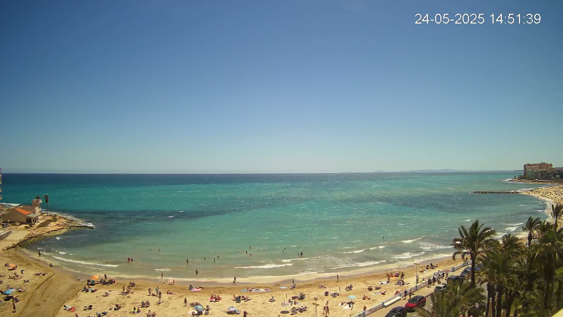 A sunny day at a sandy beach with turquoise water, showing people swimming and sunbathing, palm trees lining the shore, and buildings visible in the distance.