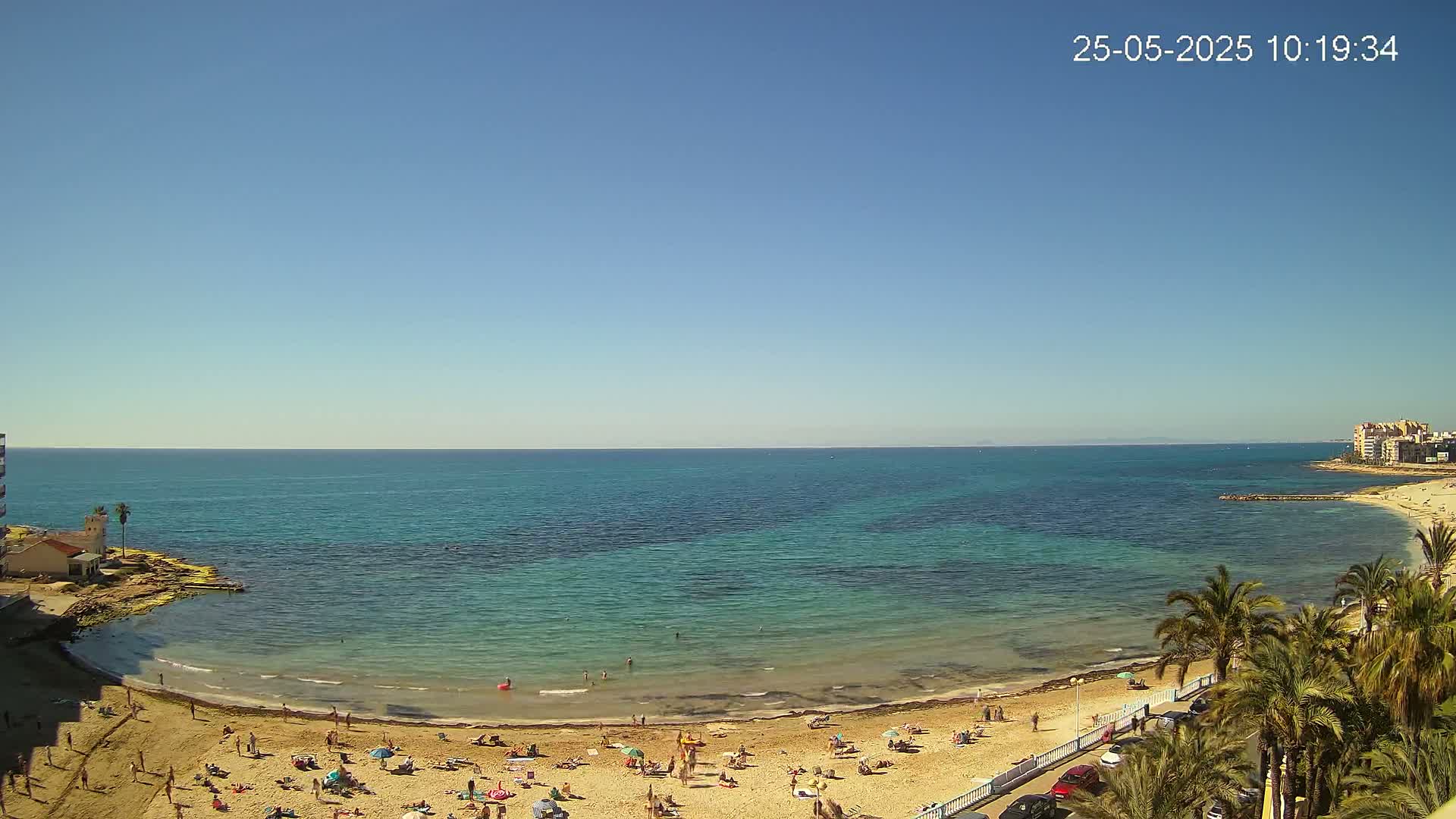 A sunny day reveals a calm, clear turquoise ocean meeting a sandy beach populated with sunbathers, palm trees lining a roadway beside the shore.