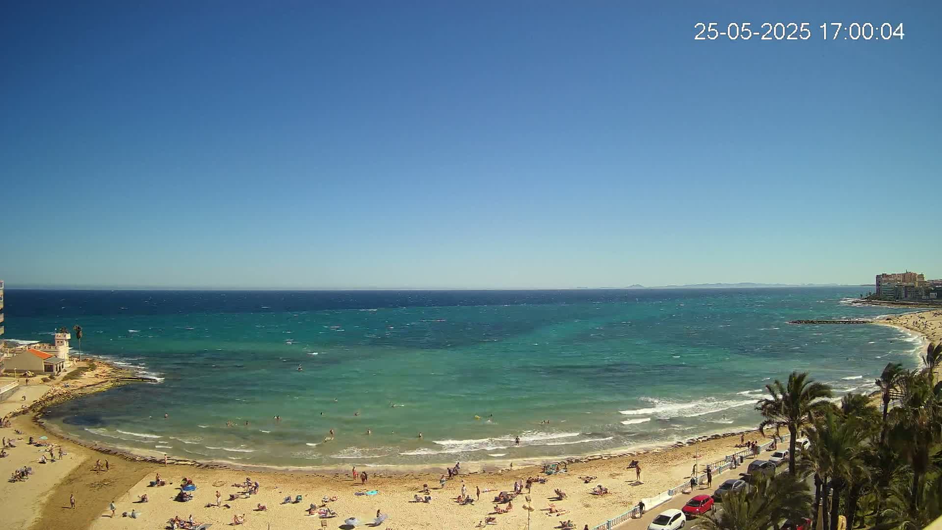 A sandy beach with bathers is situated next to a turquoise sea with small waves under a clear blue sky.