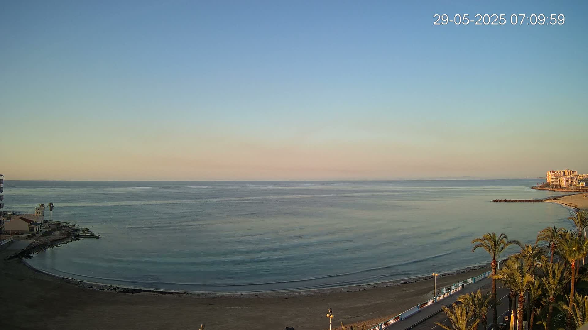 A calm, clear morning scene shows a sandy beach curving around a calm, teal ocean, with palm trees lining a road in the foreground and buildings visible in the distance.