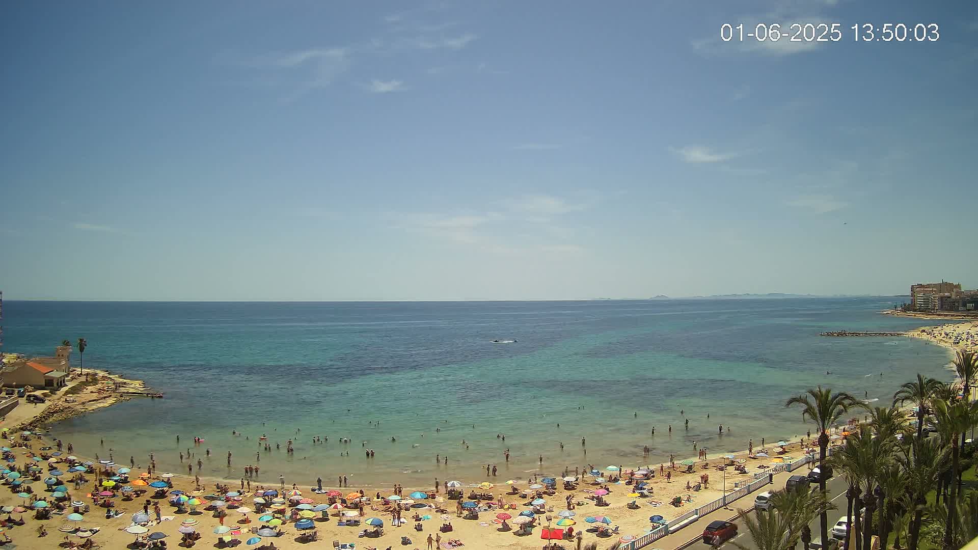 A sunny day at a crowded beach with clear, shallow turquoise water, many people swimming and sunbathing under colorful umbrellas.