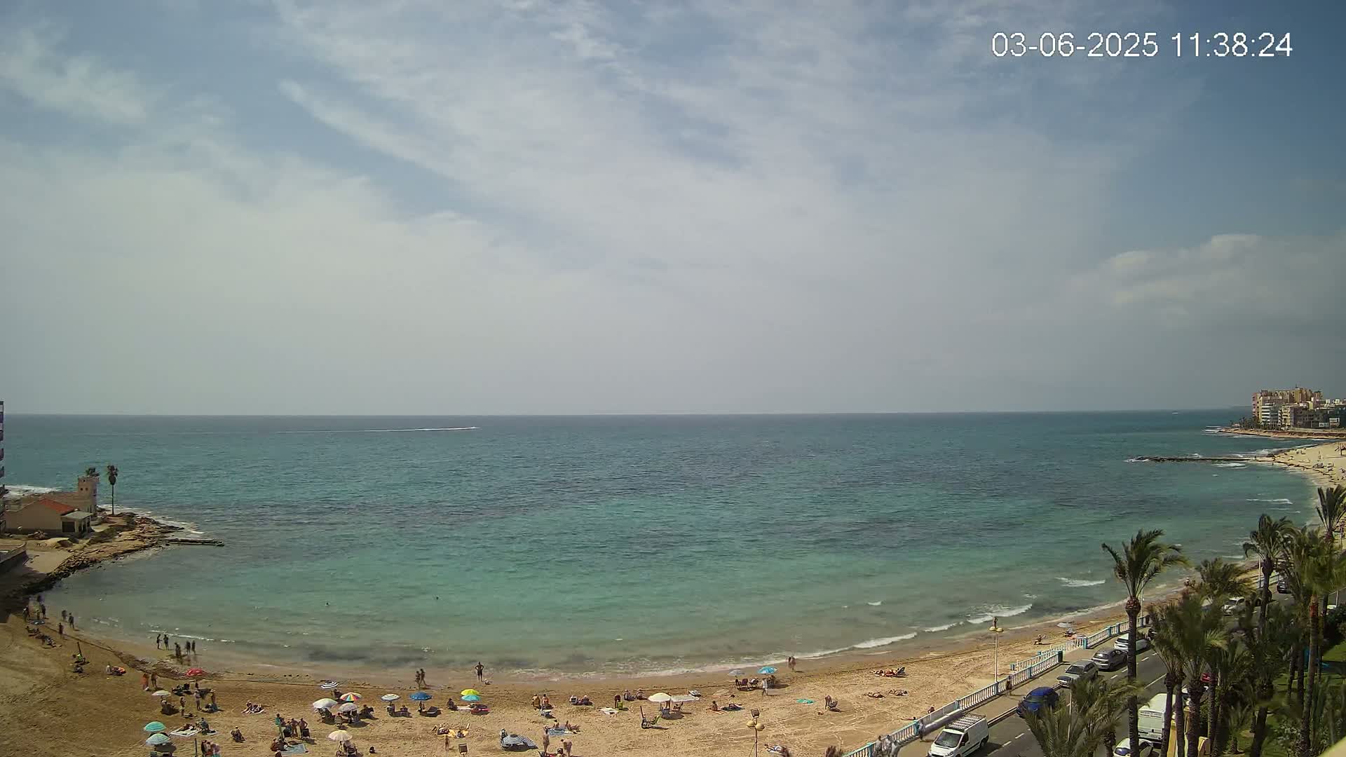 A partly cloudy day shows a sandy beach with scattered sunbathers, palm trees lining a road beside the shore, and a calm, teal-colored sea extending to the horizon.