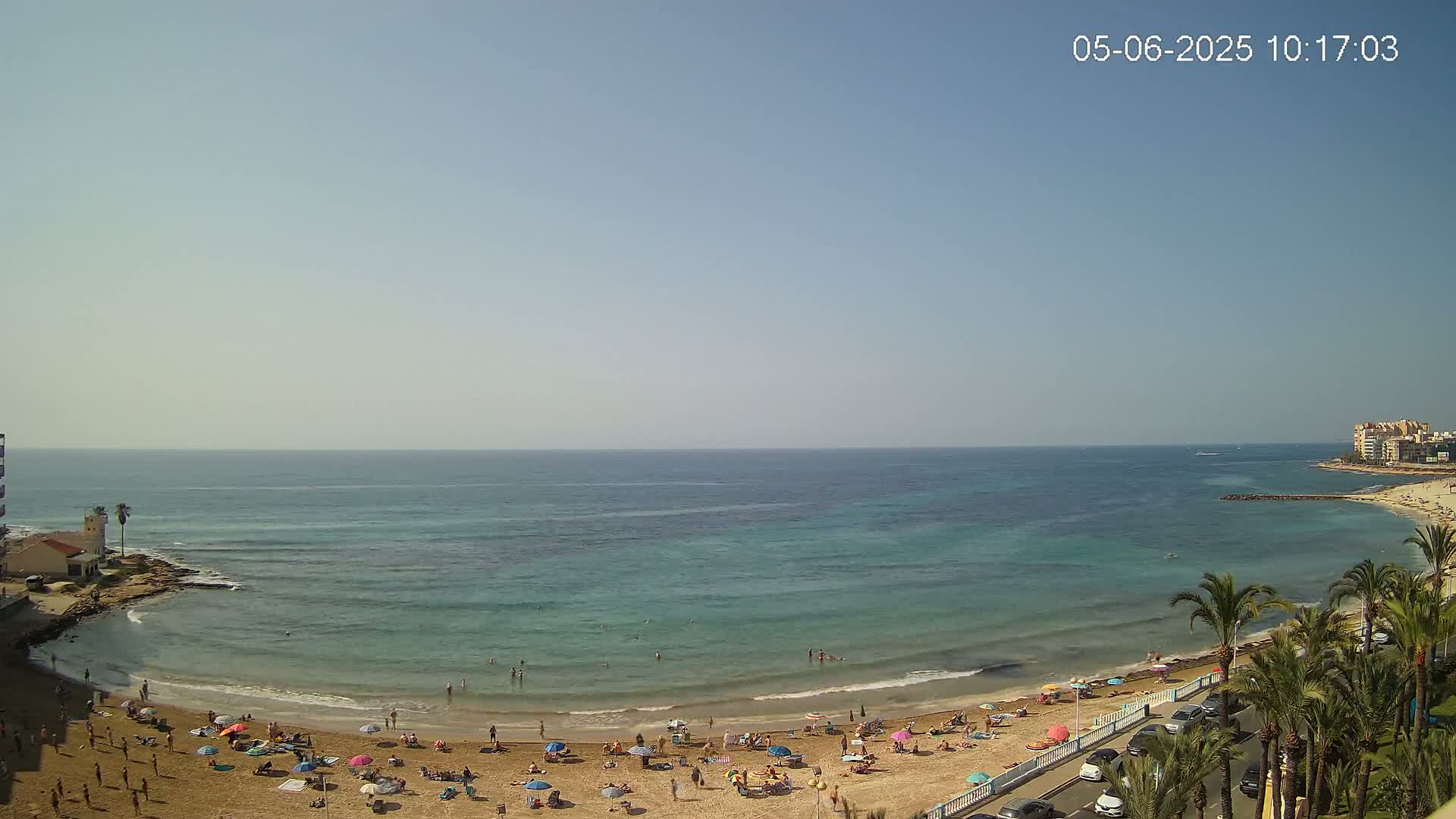 A sandy beach with many people enjoying the sunny day next to a calm, clear turquoise sea, bordered by palm trees and buildings.