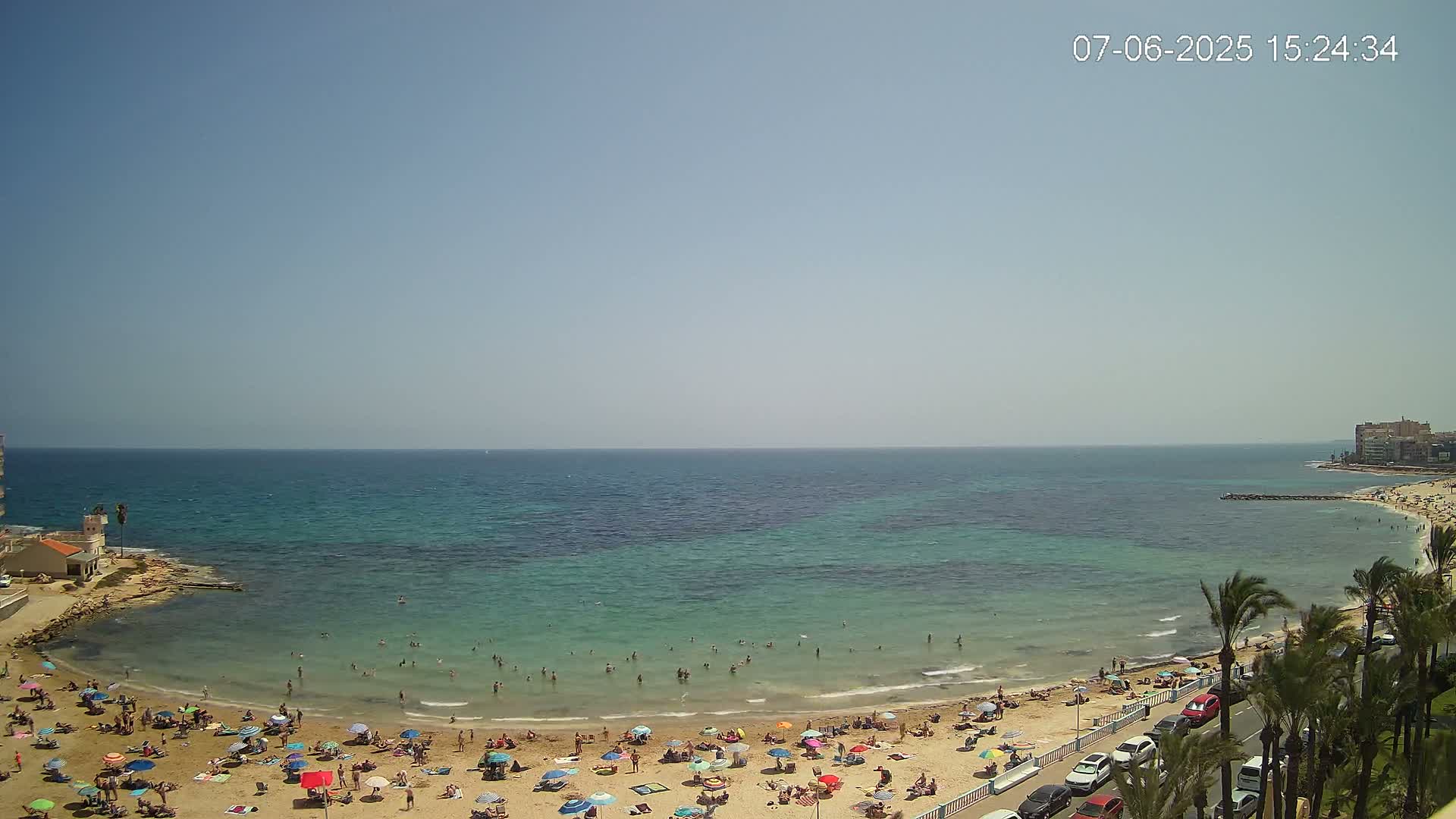 A sunny day at a beach with clear, teal water, many people swimming and sunbathing on the sand with umbrellas, and cars parked along a road lined with palm trees.