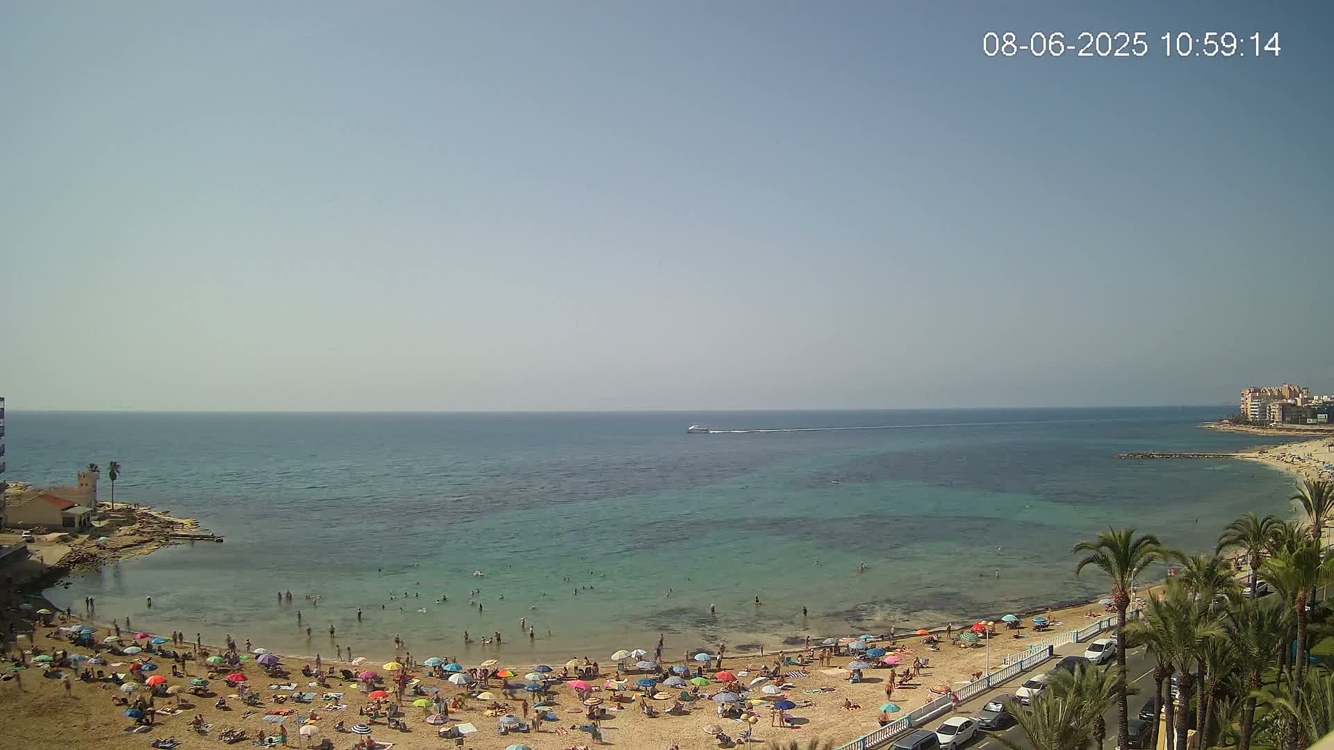 A sandy beach crowded with people under colorful umbrellas, bordered by a calm, clear turquoise sea on a sunny day.