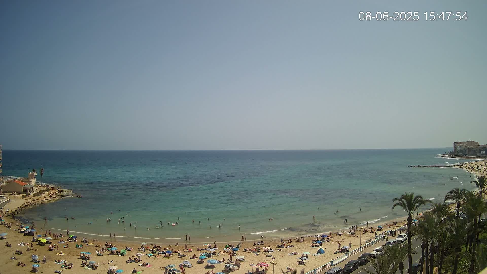 A sunny day at a beach shows a calm, teal-colored sea, a sandy shore crowded with sunbathers and swimmers, and palm trees lining a nearby road.