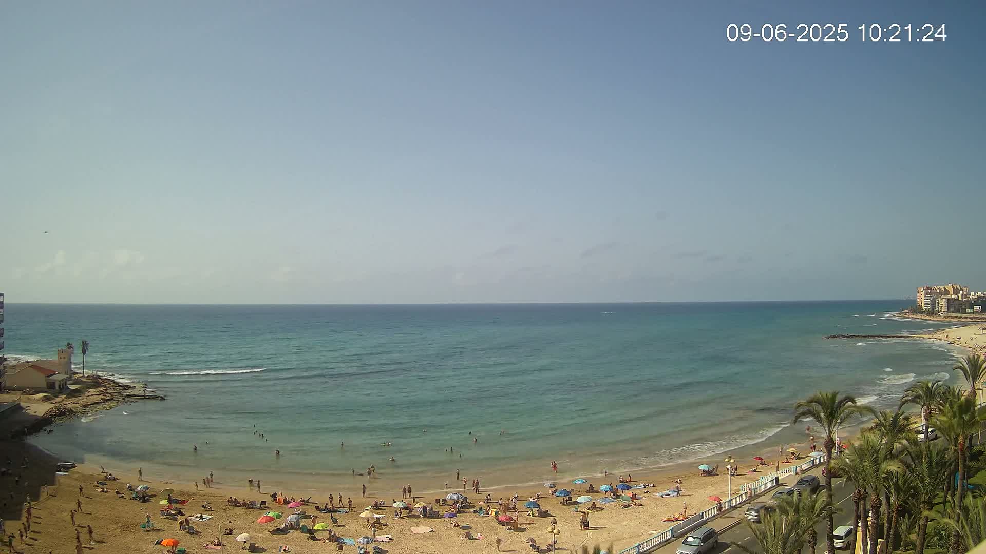 A sandy beach with numerous people and colorful umbrellas is situated along a calm, teal ocean under a clear, sunny sky.