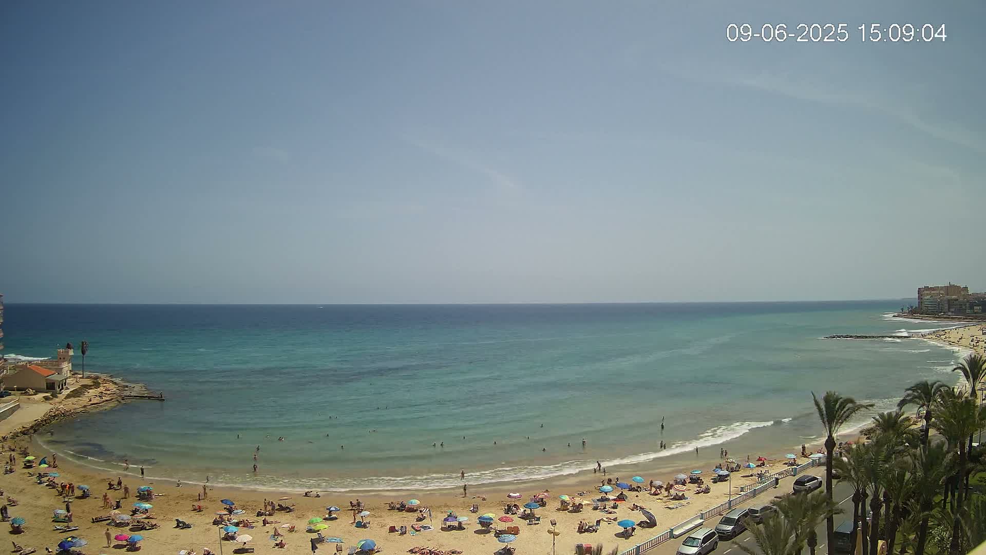 A sunny day at a beach shows a calm, turquoise sea with many people swimming and sunbathing under colorful umbrellas.