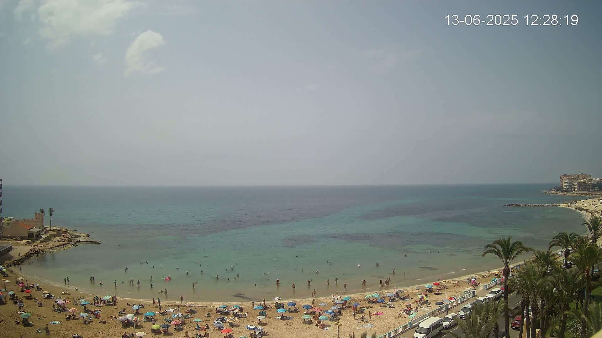 A sandy beach with numerous people and colorful umbrellas under a partly cloudy sky, next to a calm, shallow turquoise sea.