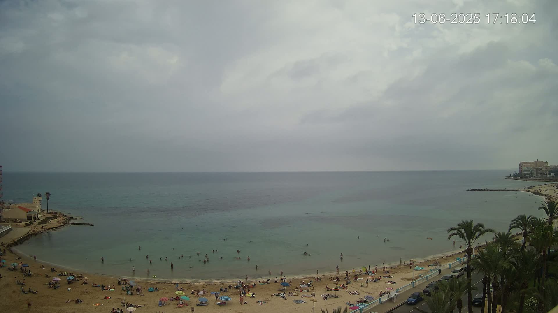 A mostly cloudy day reveals a sandy beach with many people sunbathing and swimming in calm, teal water, bordered by palm trees and buildings.