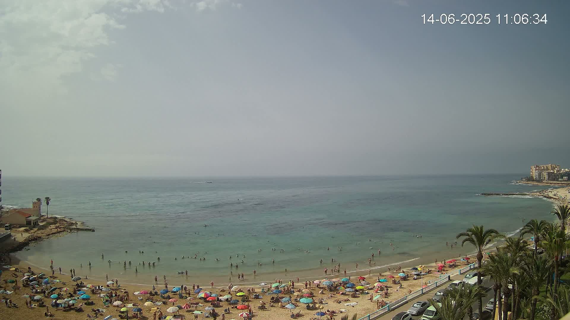 A mostly sunny day at a beach with calm, teal water, many people swimming and sunbathing under colorful umbrellas, and palm trees lining the shore.