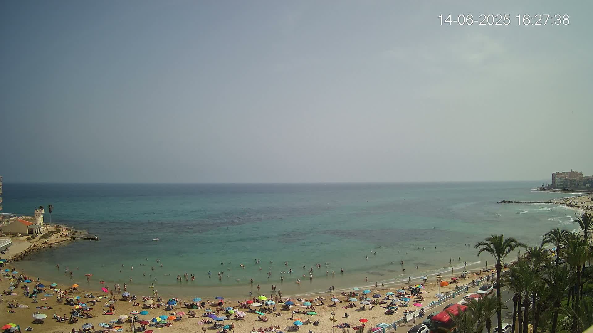 A sandy beach crowded with people under colorful umbrellas borders a calm, turquoise sea on a sunny day.