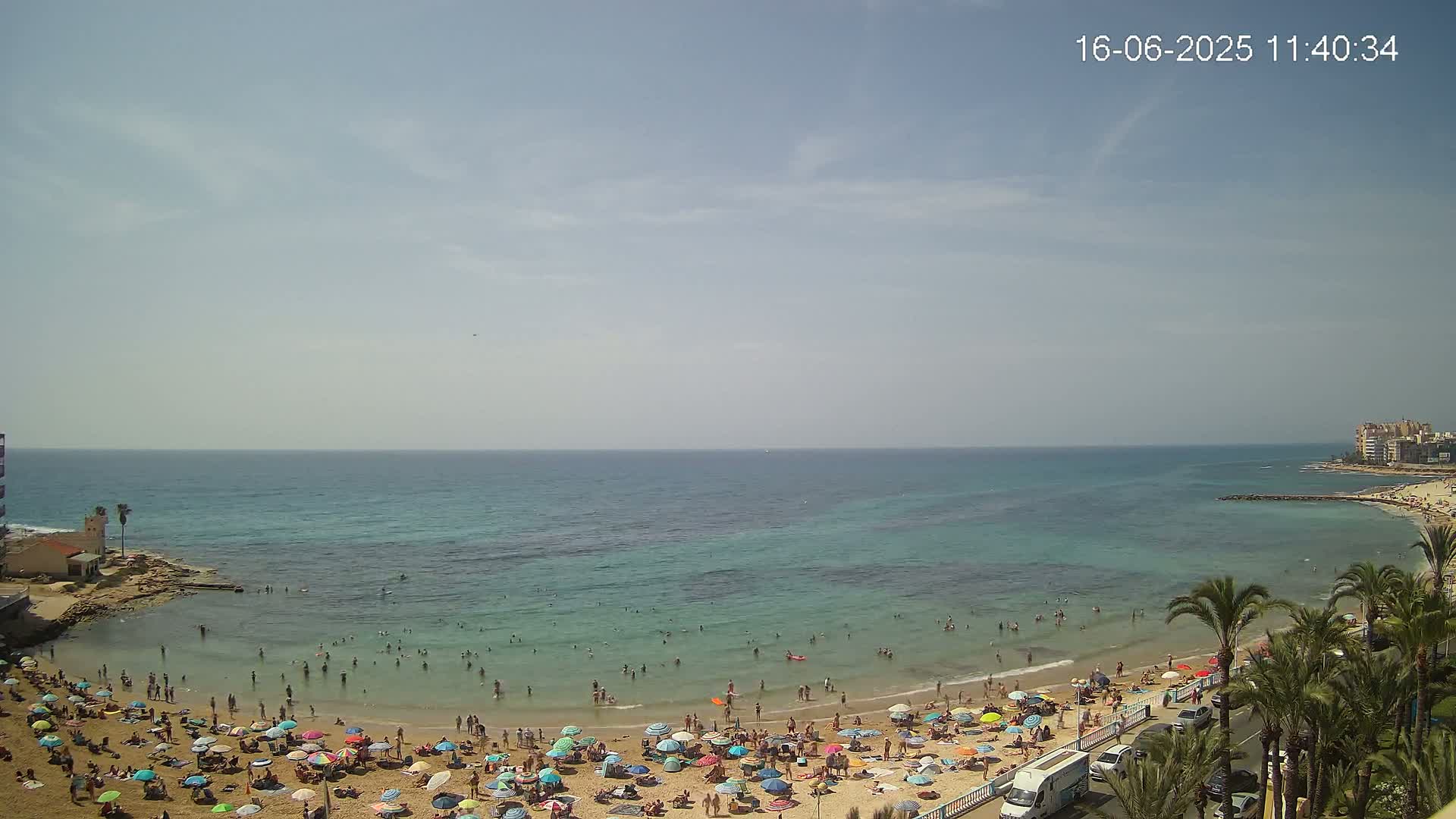 A sandy beach crowded with people under colorful umbrellas, next to a calm, clear turquoise ocean under a mostly sunny sky.