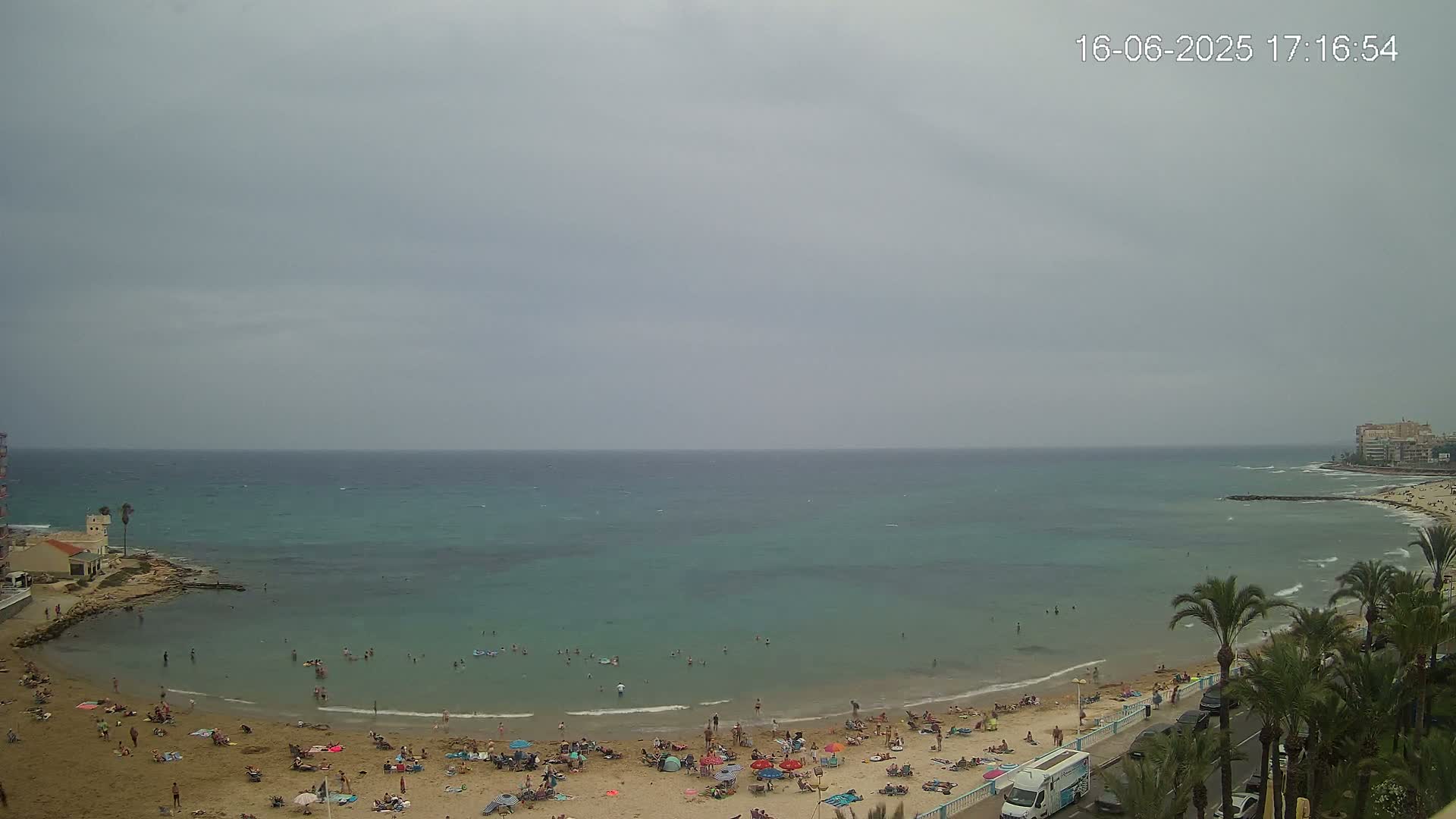A beach with many people on it is situated next to a calm, teal-colored sea under an overcast sky.