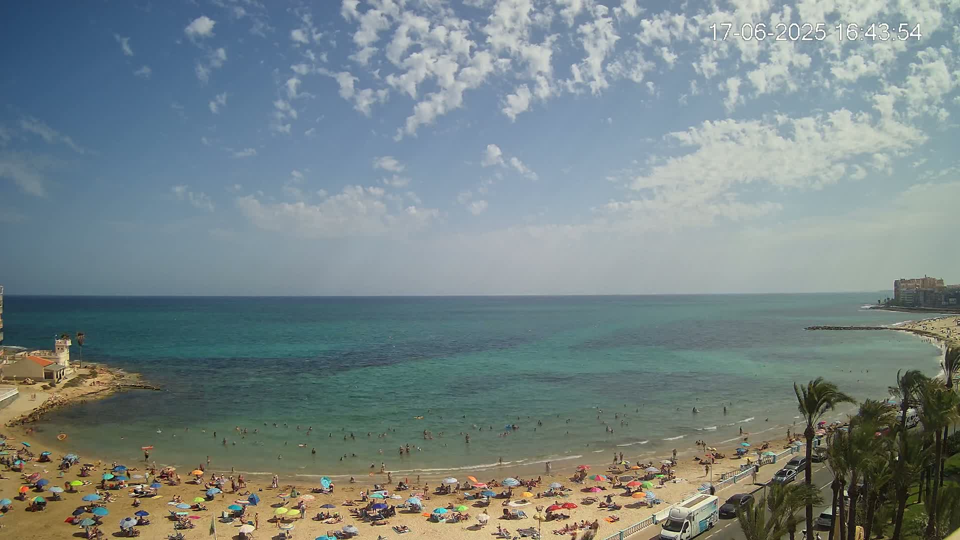 A partly cloudy day at a beach with turquoise water, where many people are swimming and sunbathing under colorful umbrellas.
