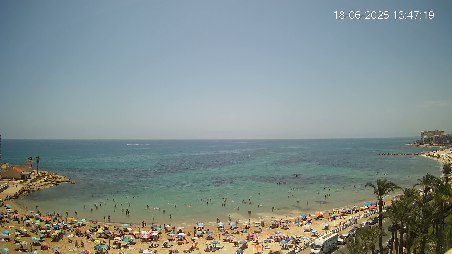 A sunny day at a crowded beach with turquoise water, numerous colorful umbrellas, and palm trees lining the shore.
