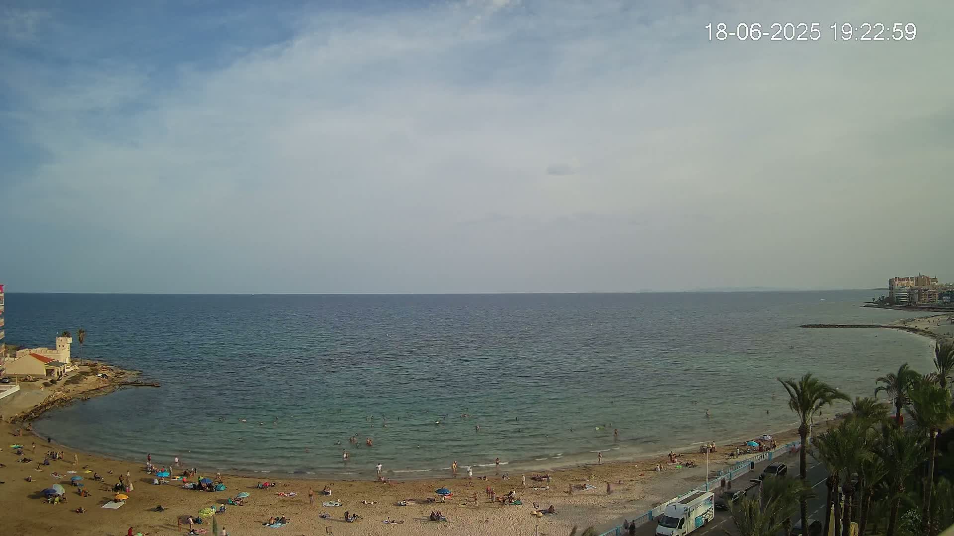 A sandy beach with people sunbathing and swimming in a calm sea under a partly cloudy sky.