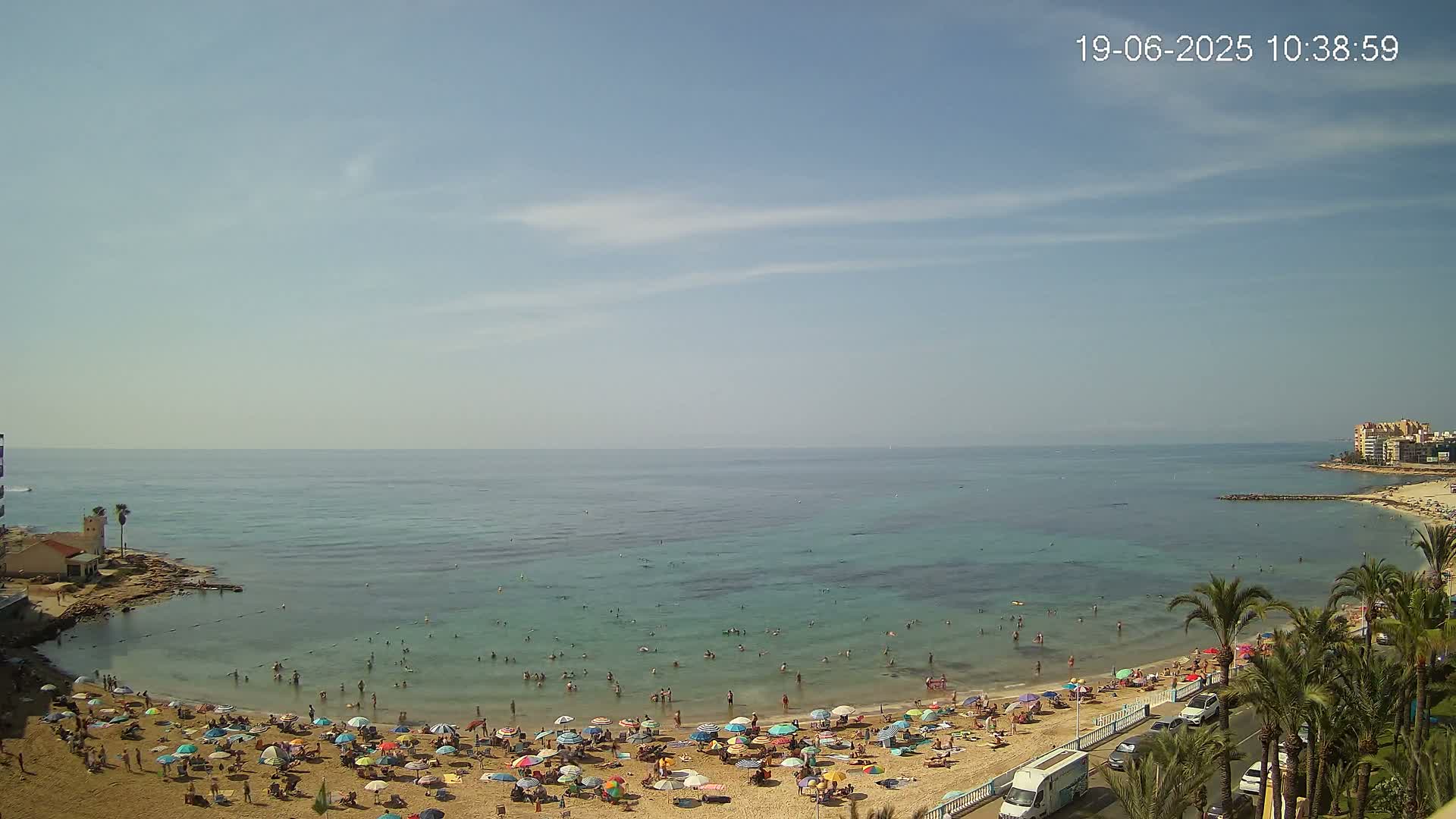 A sunny day at a sandy beach with clear, calm water, many people swimming and sunbathing under colorful umbrellas, and palm trees lining a nearby road.
