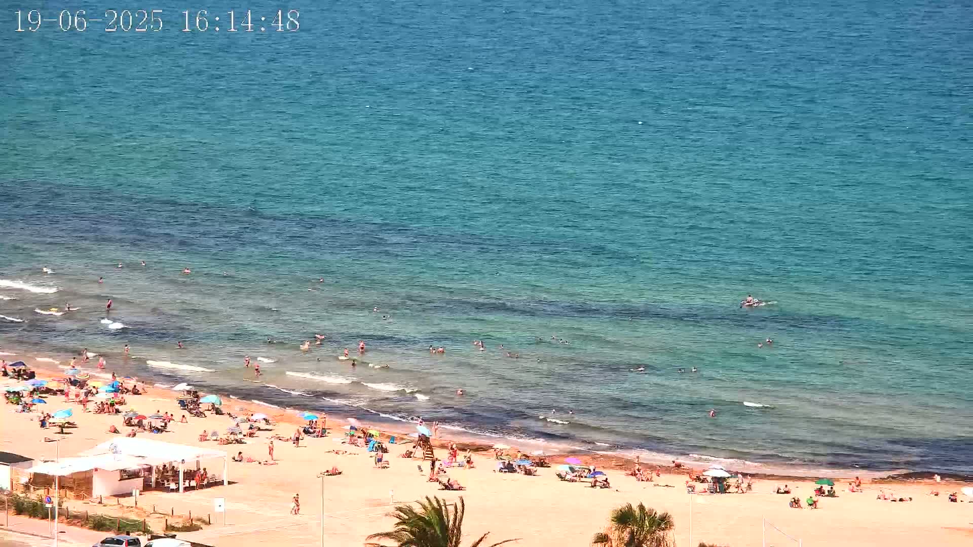 A sandy beach with many people swimming and sunbathing, under a clear sunny sky, next to a calm, clear blue ocean.