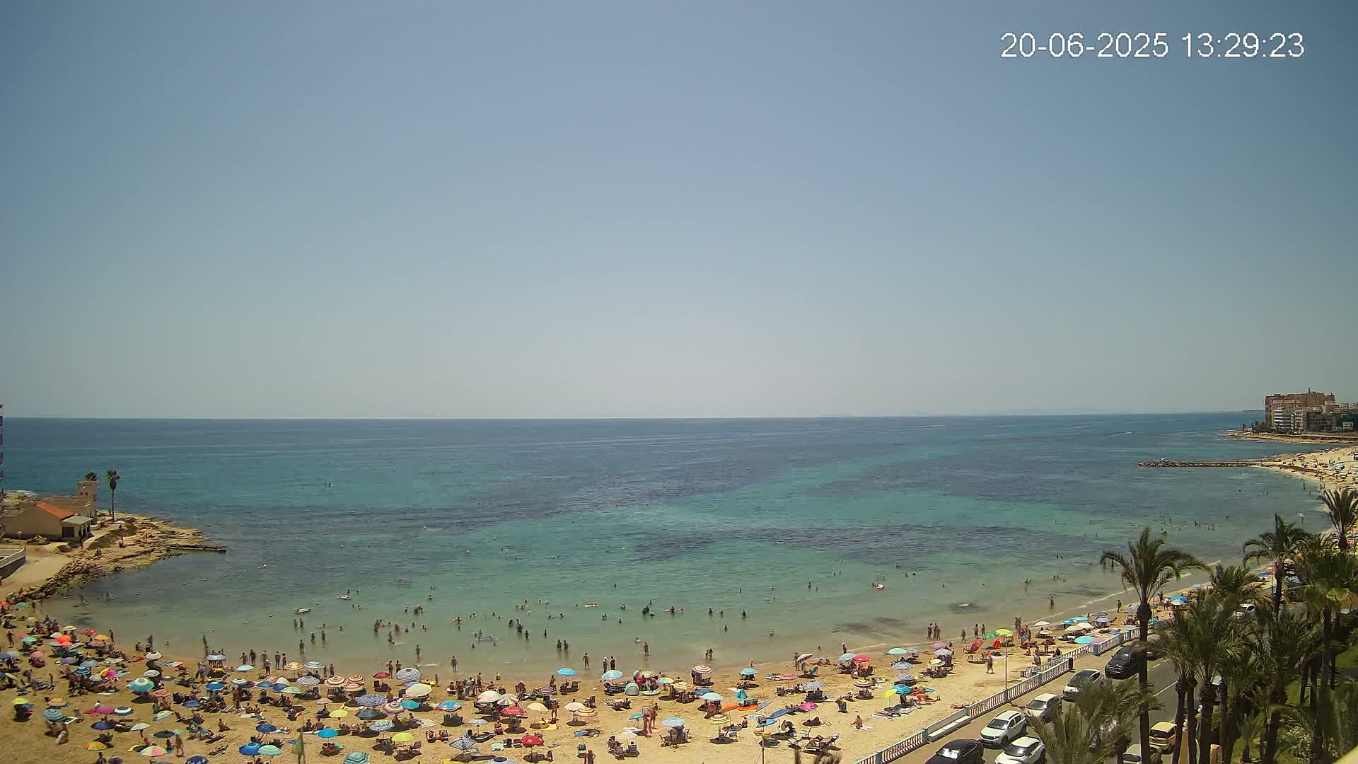 A sunny day at a crowded beach with clear, teal water and numerous beach umbrellas.