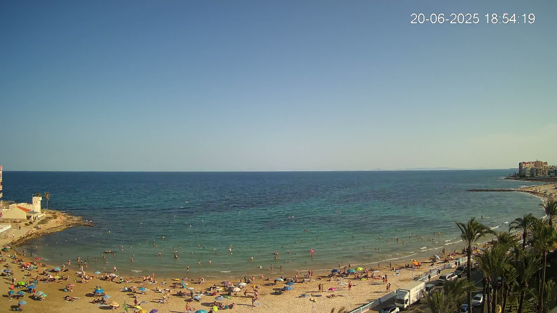 A sunny day at a sandy beach shows many people swimming and sunbathing under colorful umbrellas, with palm trees and buildings visible in the background.