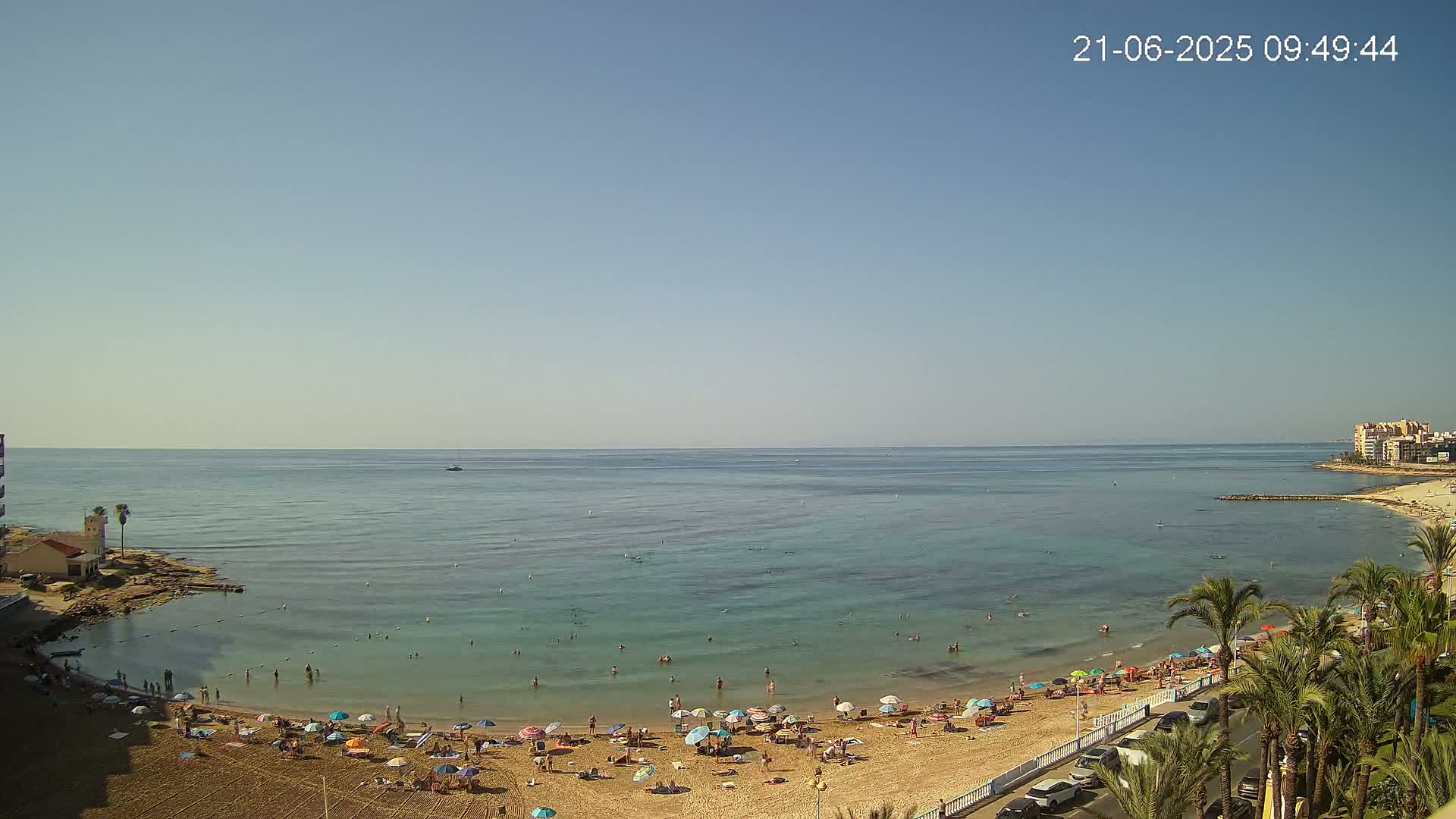A sandy beach with numerous umbrellas and people is situated next to calm, clear turquoise water under a clear blue sky.