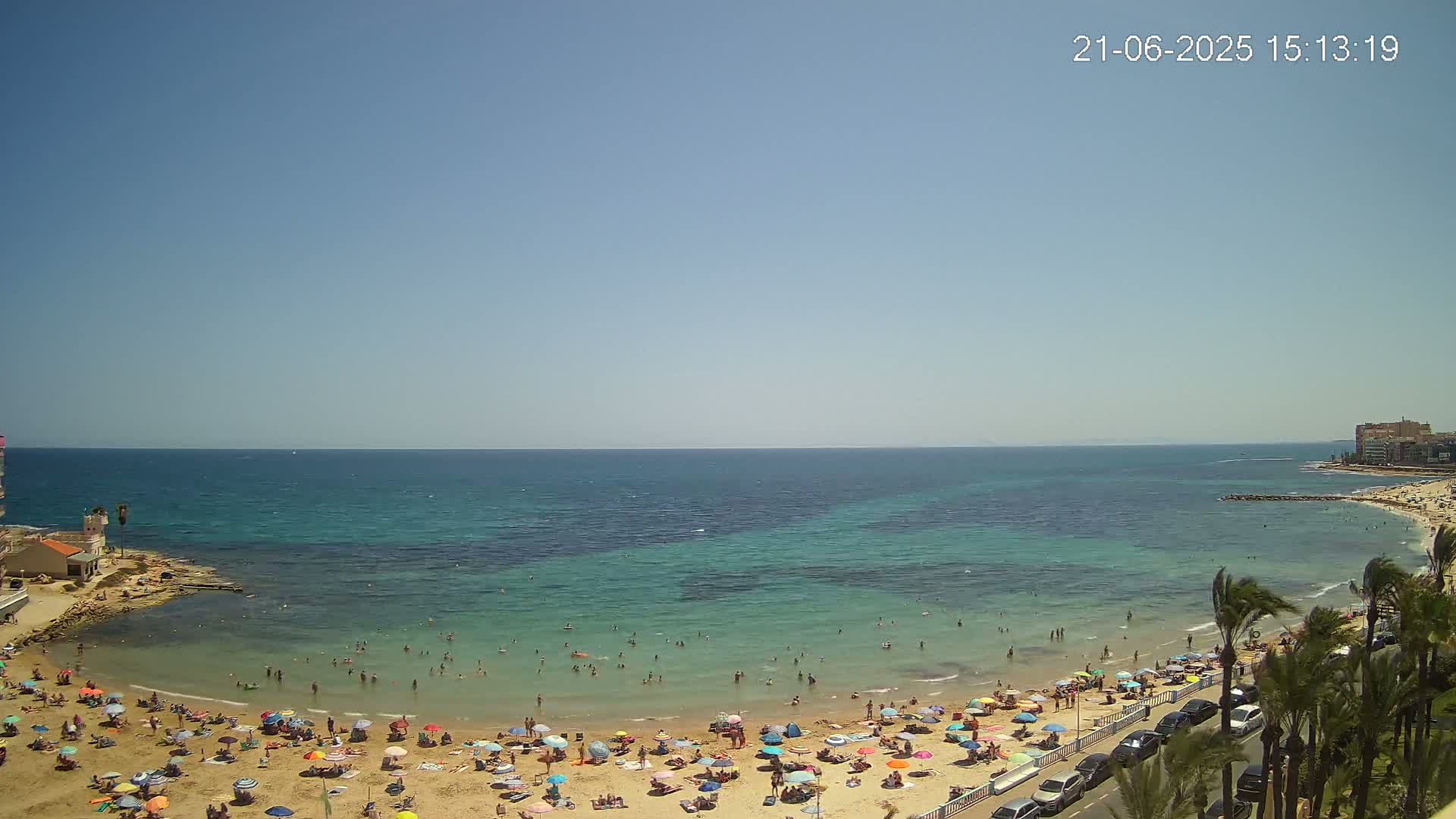 A sunny day at a sandy beach with calm, clear turquoise water, many people swimming and sunbathing under colorful umbrellas, and palm trees lining a road nearby.