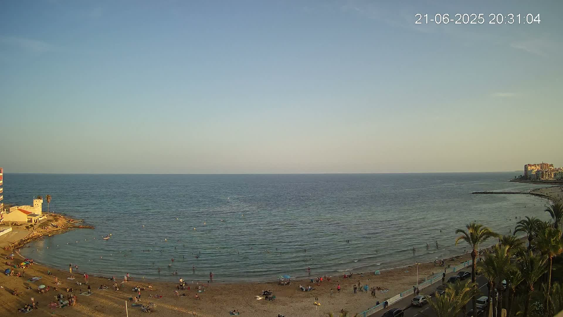 A sandy beach with people swimming and sunbathing is bordered by palm trees and buildings under a clear, pale sky at dusk.