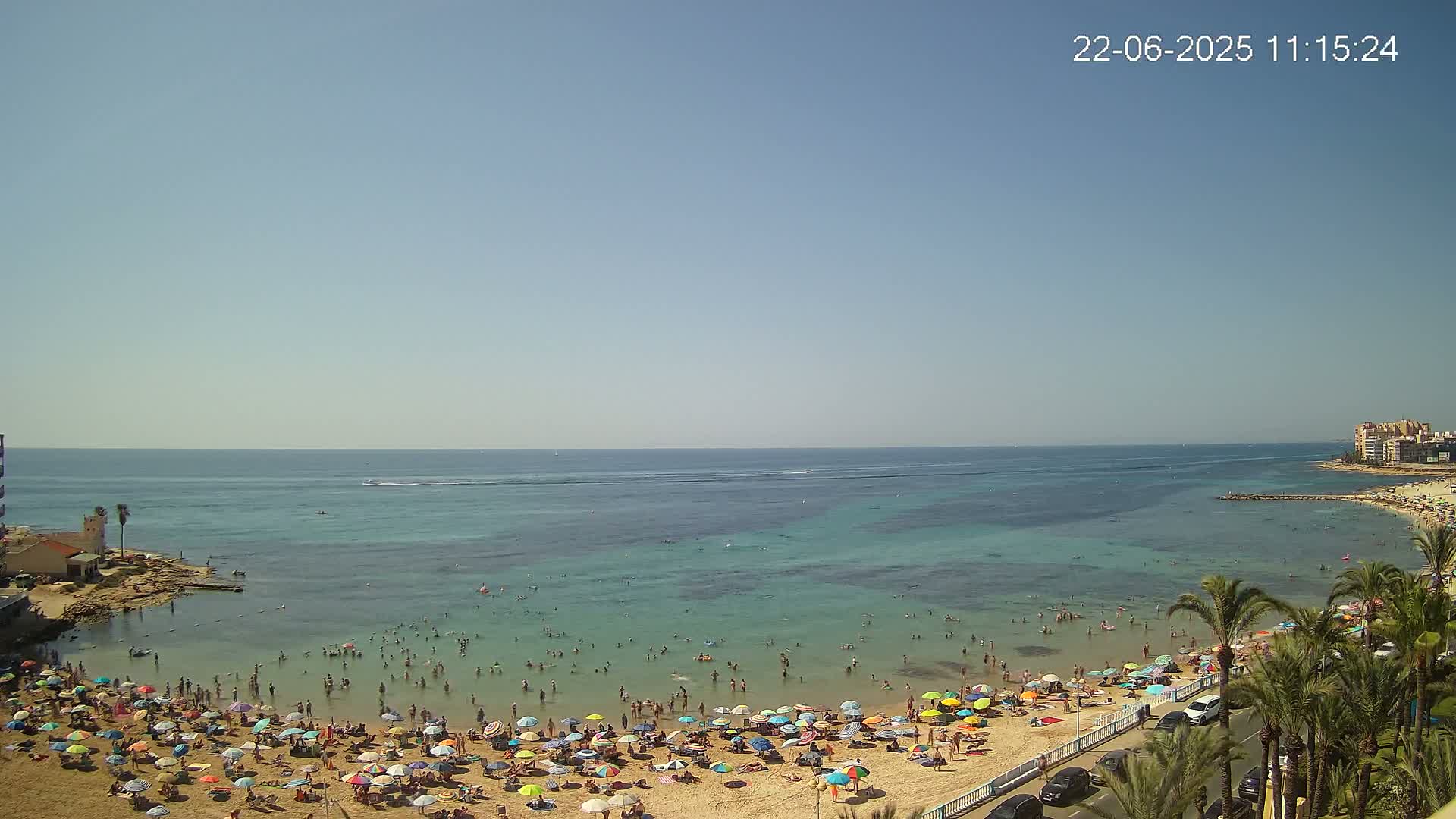 A crowded sandy beach with many colorful umbrellas under a clear, sunny sky, next to calm, shallow turquoise water.
