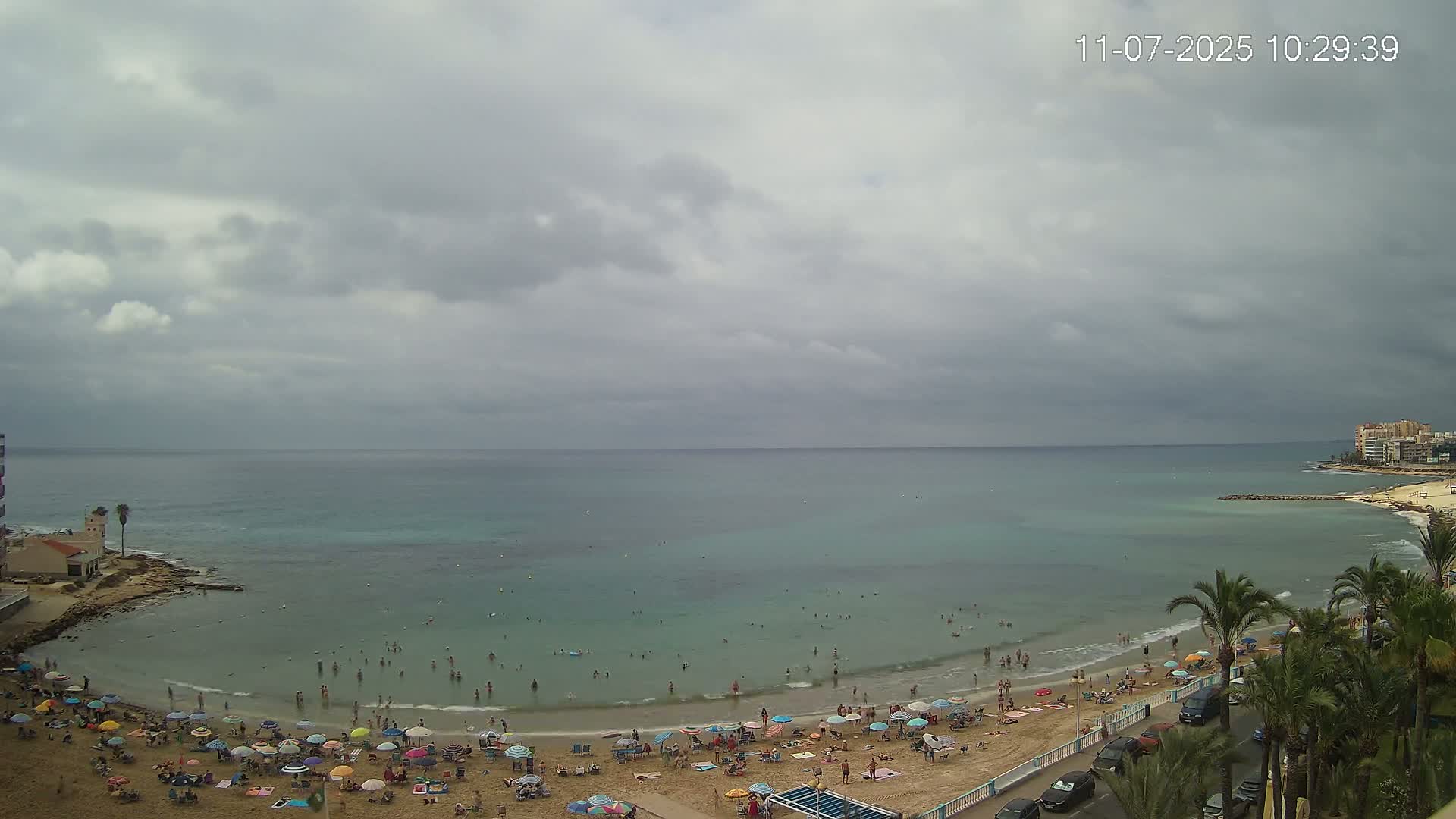 A sandy beach with many colorful umbrellas and people swimming in calm, teal water under an overcast sky.