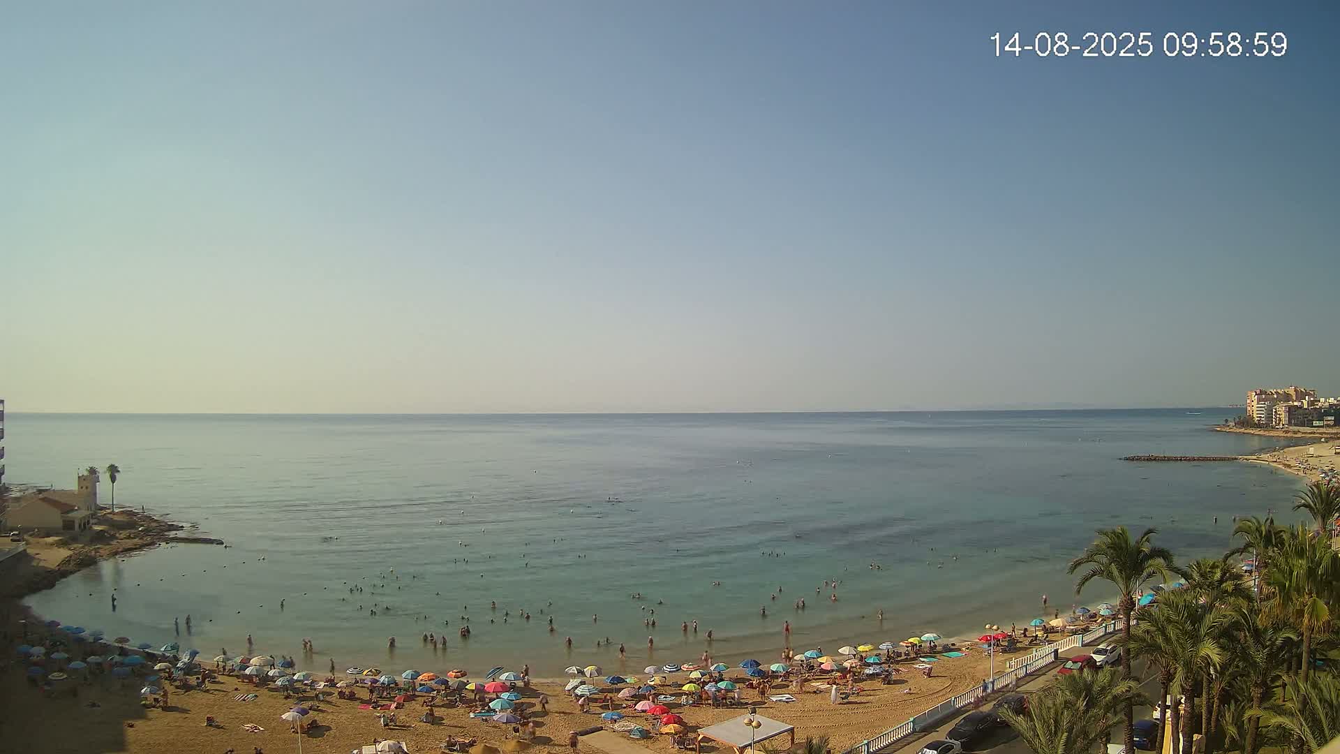 A sunny day at a beach shows many people swimming and relaxing under colorful umbrellas on a sandy shore, with calm, clear water extending to a distant coastline.