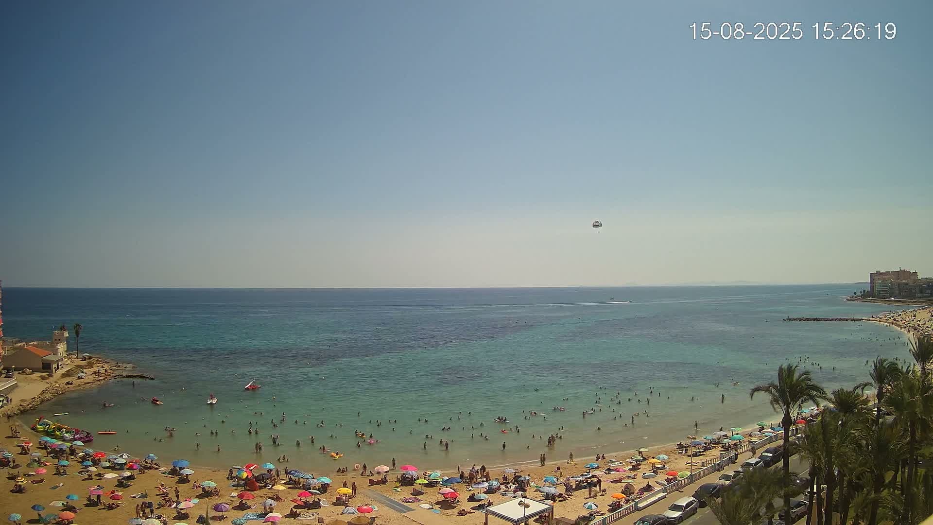 A sunny day at a beach shows many people swimming and relaxing under colorful umbrellas on a sandy shore, with calm, clear water extending to a distant coastline.