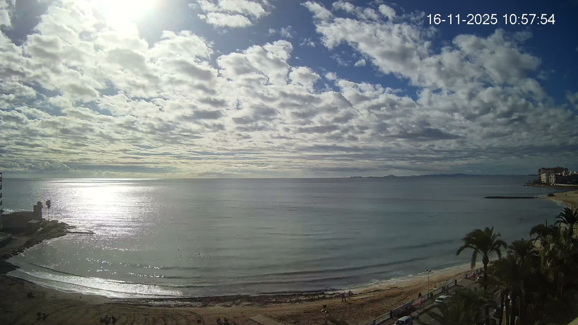 A sunny, partly cloudy day overlooks a wide, calm ocean reflecting sunlight, bordered by a sandy beach with a few scattered people and coastal buildings with palm trees on either side.