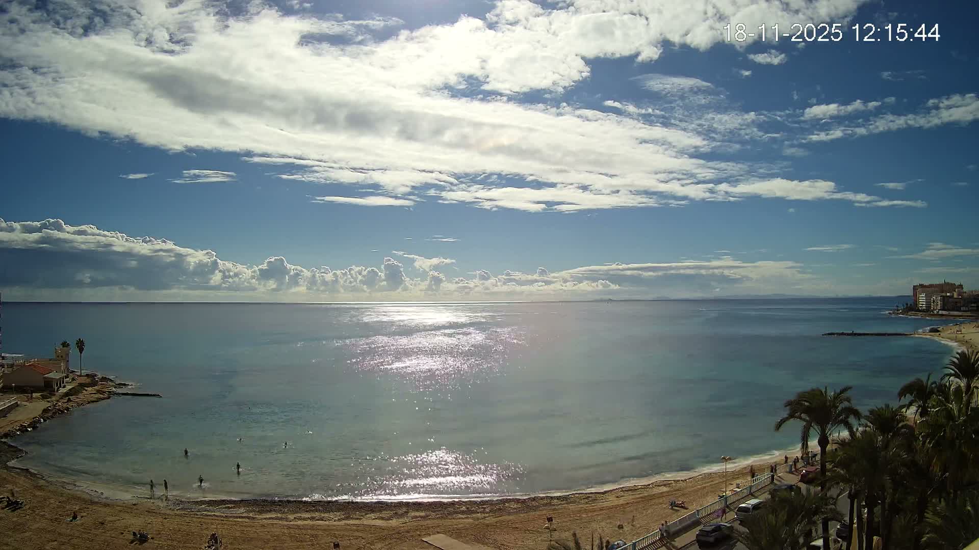 A sunny, partly cloudy day overlooks a wide, calm ocean reflecting sunlight, bordered by a sandy beach with a few scattered people and coastal buildings with palm trees on either side.