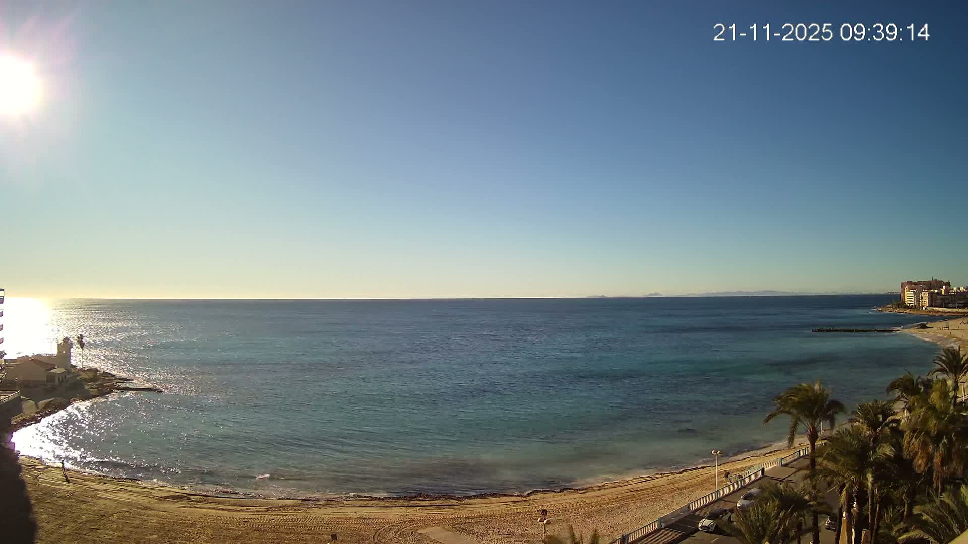 A bright sunny day illuminates a wide coastal scene featuring a sandy beach, calm turquoise-blue ocean, a prominent lighthouse on the left shore, distant buildings and palm trees lining a promenade on the right, all under a clear blue sky.
