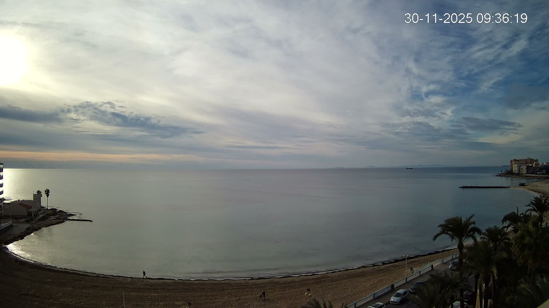 A panoramic view captures a calm sea meeting sandy beaches where a few people stroll, flanked by coastal buildings and palm trees under a partly cloudy sky with morning sunlight breaking through on the left.