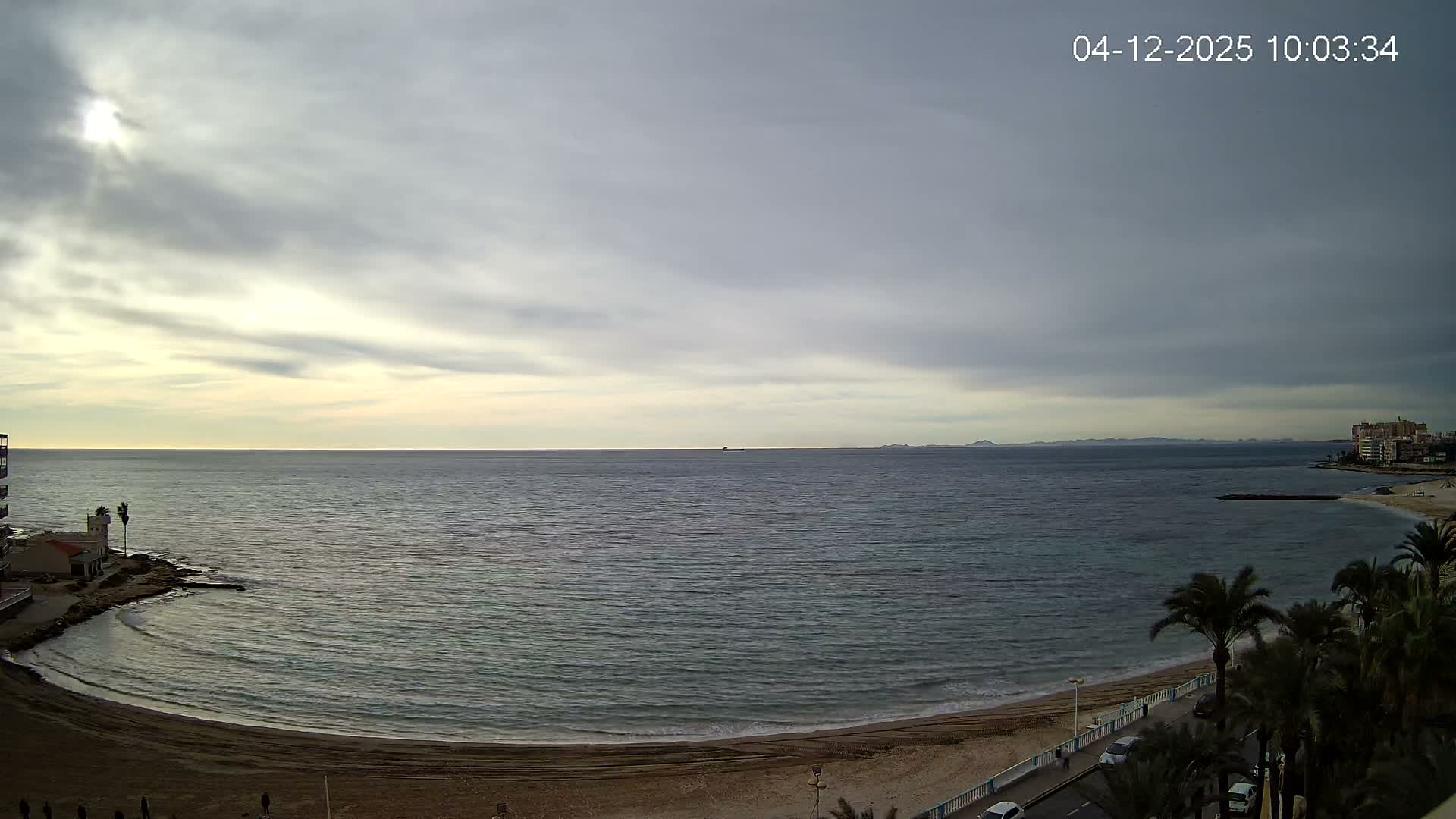 A wide coastal view under a mostly cloudy sky with bright sunlight breaking through on the left, showing a calm ocean, a sandy beach, a promenade with palm trees and coastal buildings, and a distant ship on the horizon.