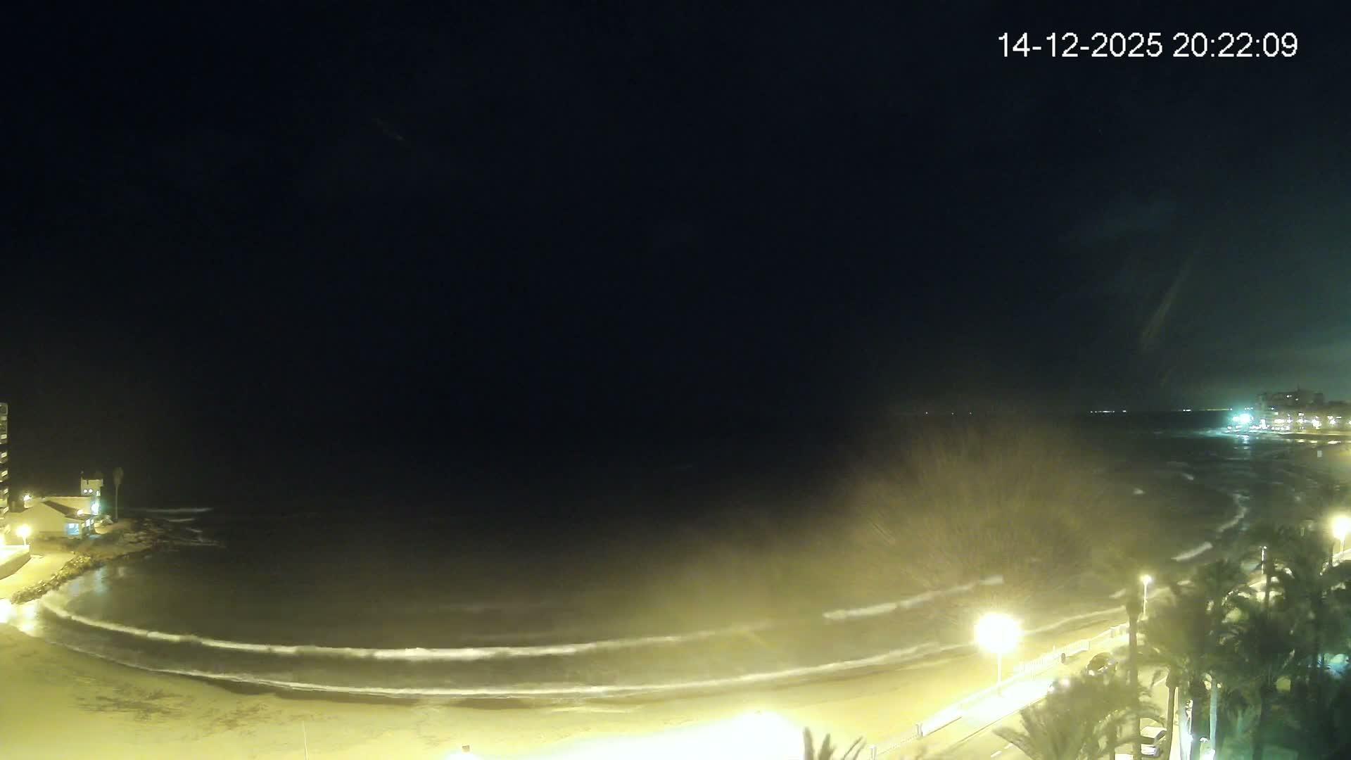 A wide coastal view under a mostly cloudy sky with bright sunlight breaking through on the left, showing a calm ocean, a sandy beach, a promenade with palm trees and coastal buildings, and a distant ship on the horizon.