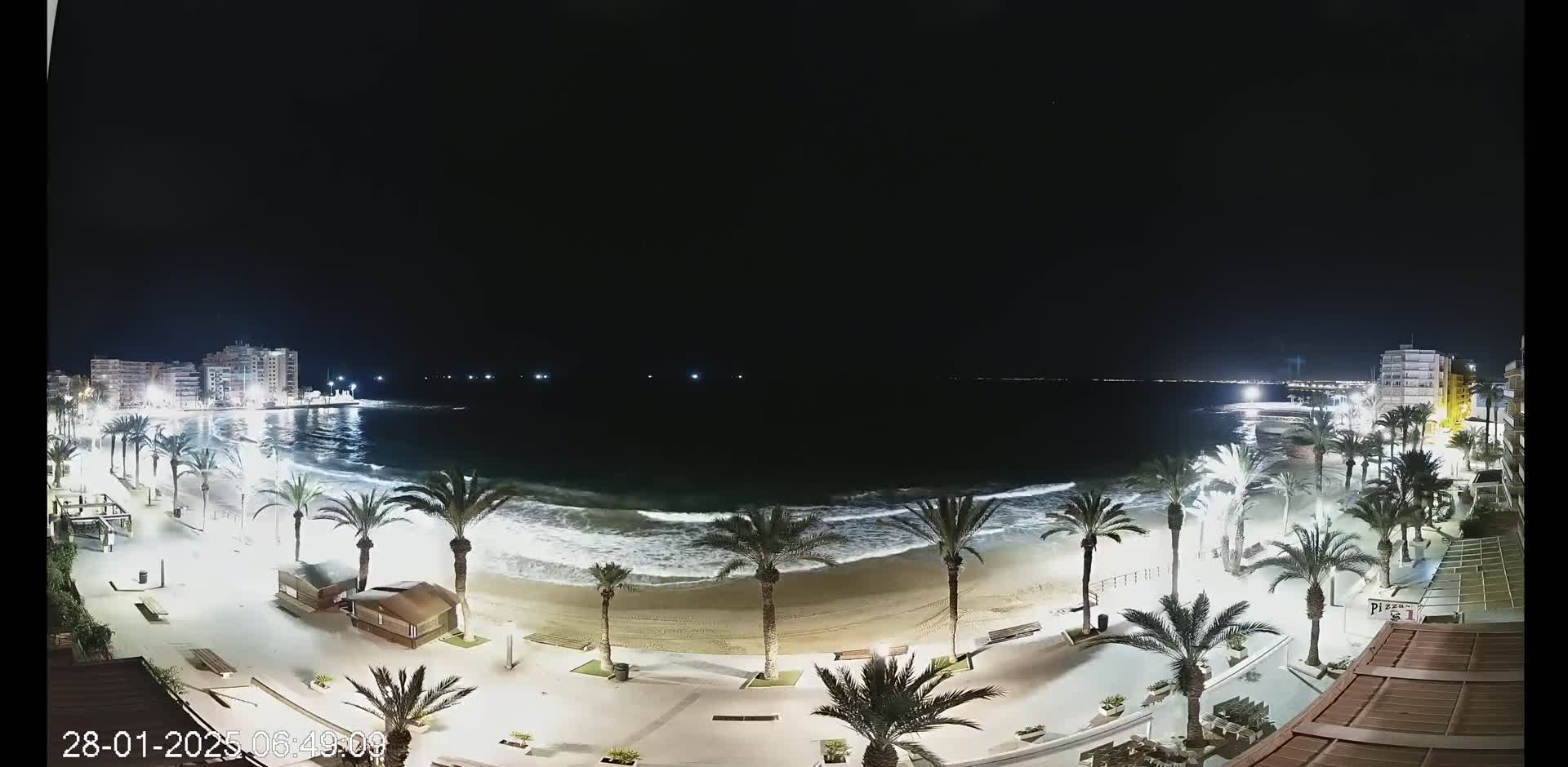 A nighttime view of a sandy beach with palm trees lining the shoreline, showing moderate waves and buildings illuminated in the distance under a clear, dark sky.