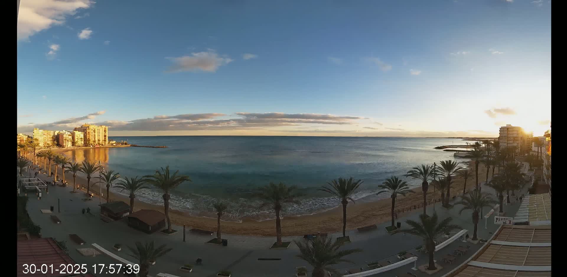 A beach with palm trees lines a seaside promenade at sunset under a mostly clear sky.