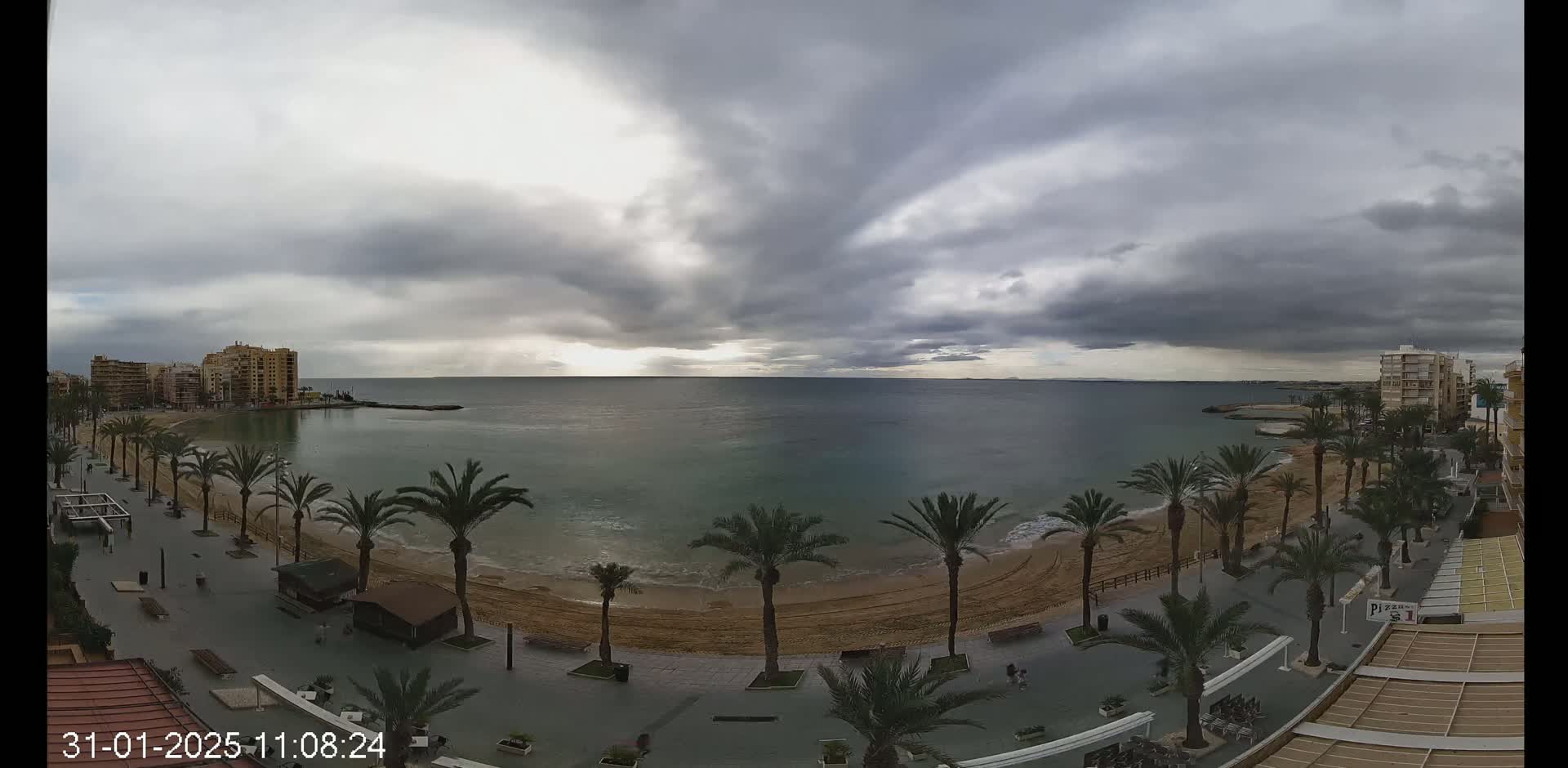 A mostly cloudy day shows a sandy beach lined with palm trees and buildings overlooking a calm sea.